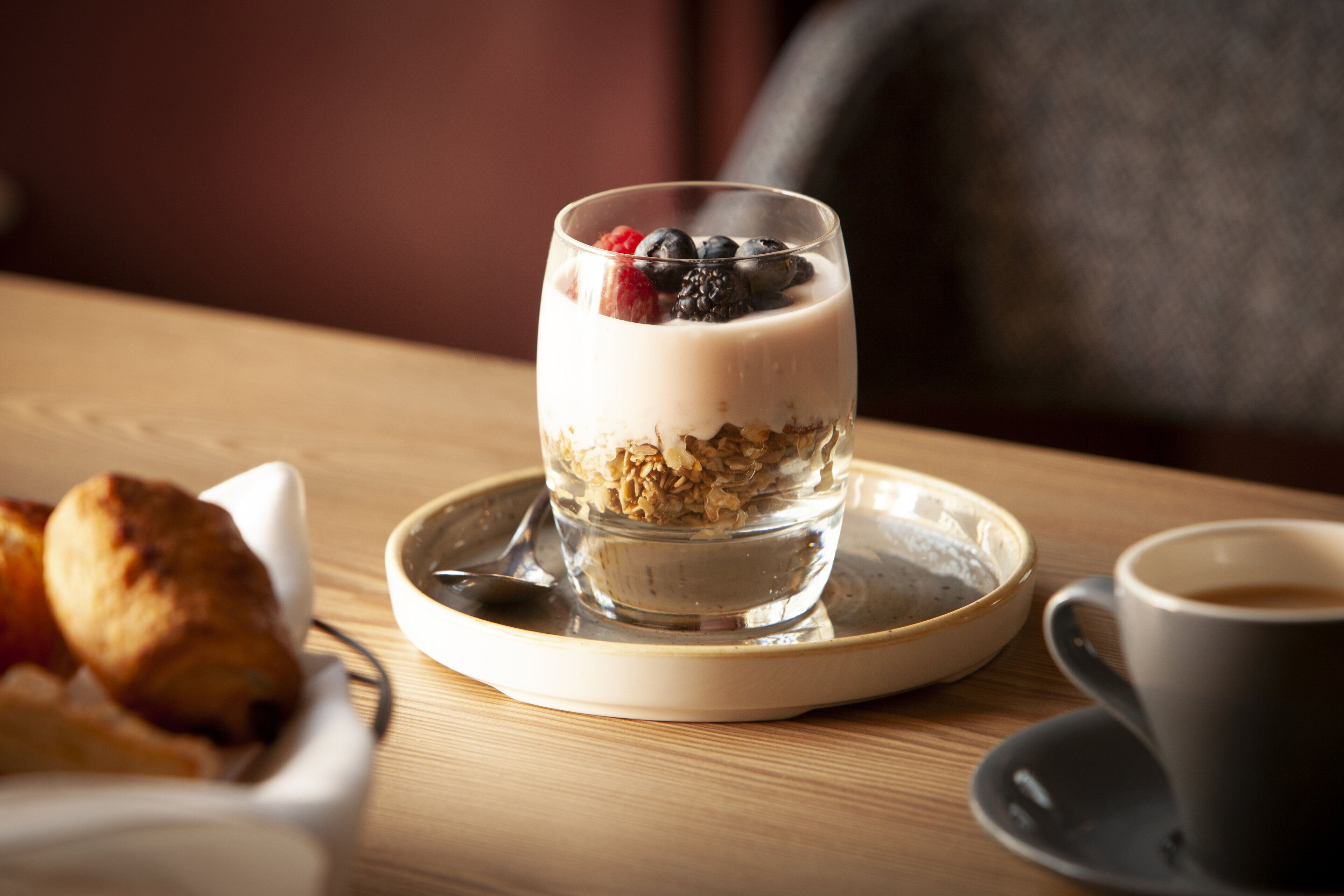 A breakfast setting with a glass of yogurt topped with berries and granola, a croissant, and a cup of coffee on a wooden table.