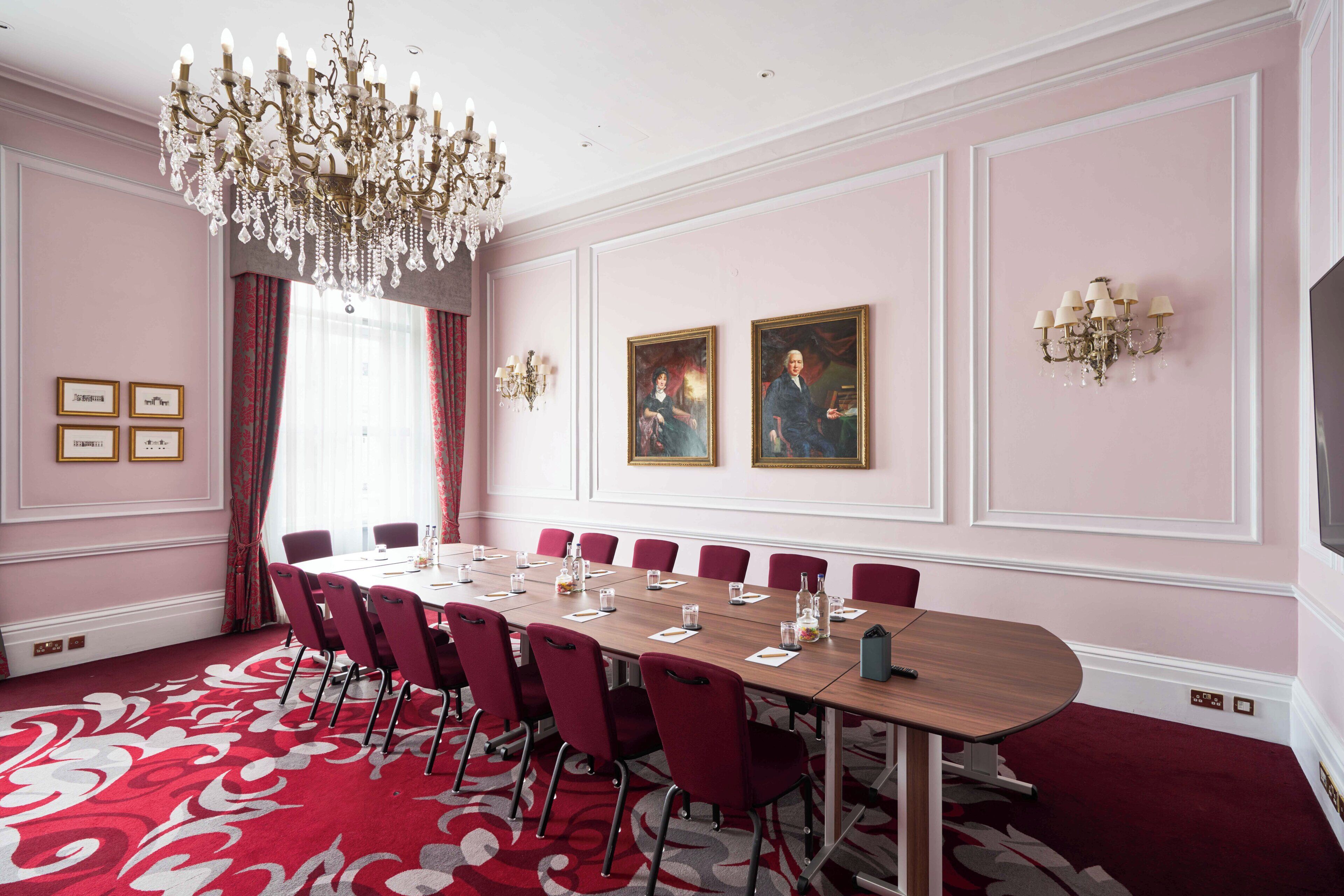 A conference room with a long table, red chairs, a chandelier, framed pictures, and a red carpet.