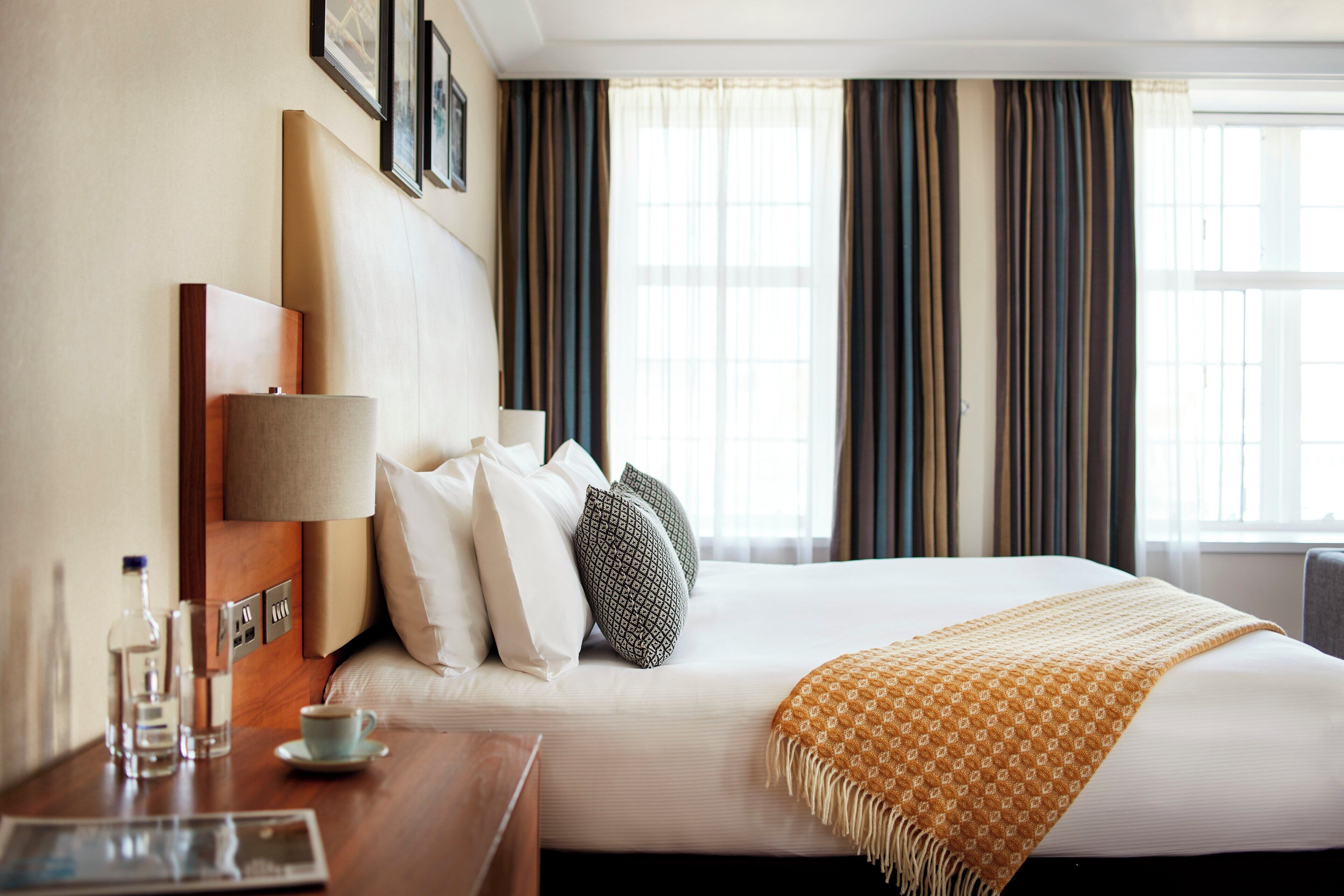 A hotel room with a neatly made bed, a bedside table with a lamp, a cup, and a newspaper.