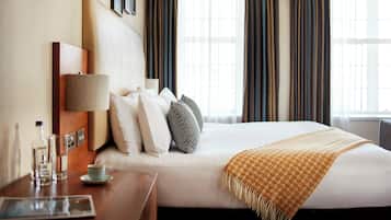 A hotel room with a neatly made bed, a bedside table with a lamp, a cup, and a newspaper.