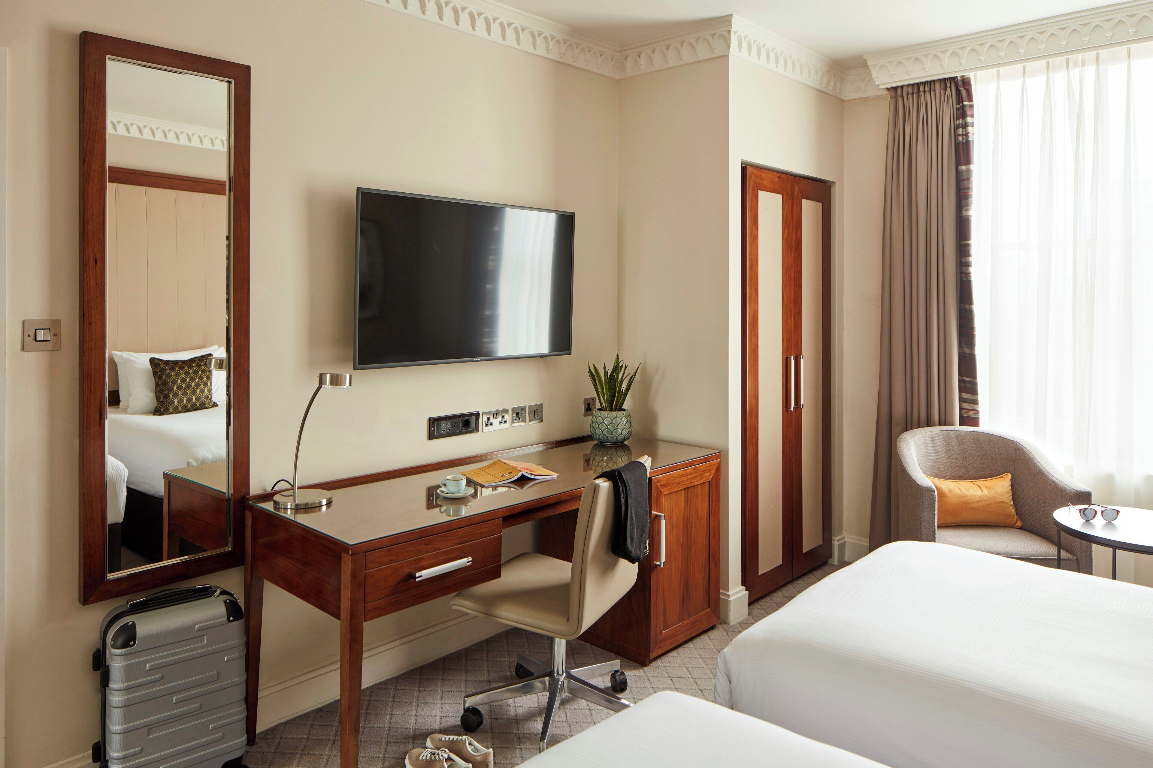A hotel room with a bed, a desk with a chair, a television, a mirror, a wardrobe, and a small table with a plant.