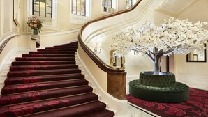 A grand staircase with a red carpet, a green ottoman, and a white artificial tree. - The Clermont London, Charing Cross (London)