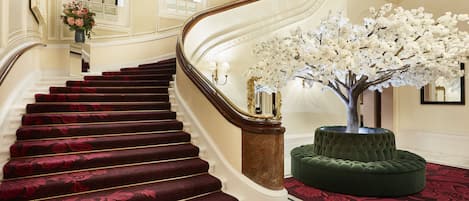 Una imponente escalera con alfombra roja, un reposapiés verde y un árbol artificial blanco.