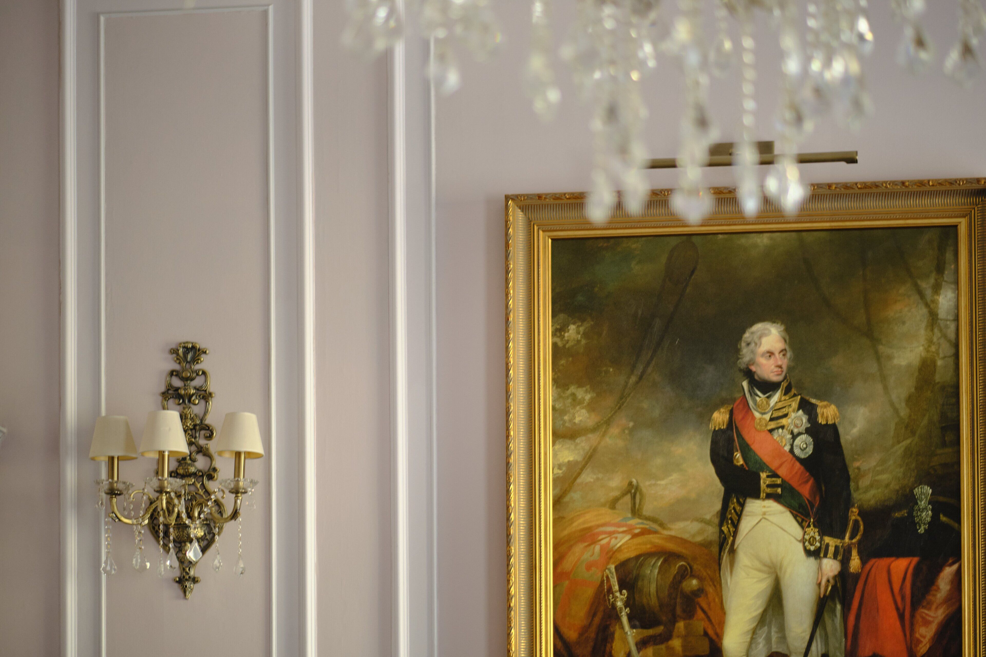 A room with a large portrait of a historical figure, a chandelier, and a wall-mounted light fixture.