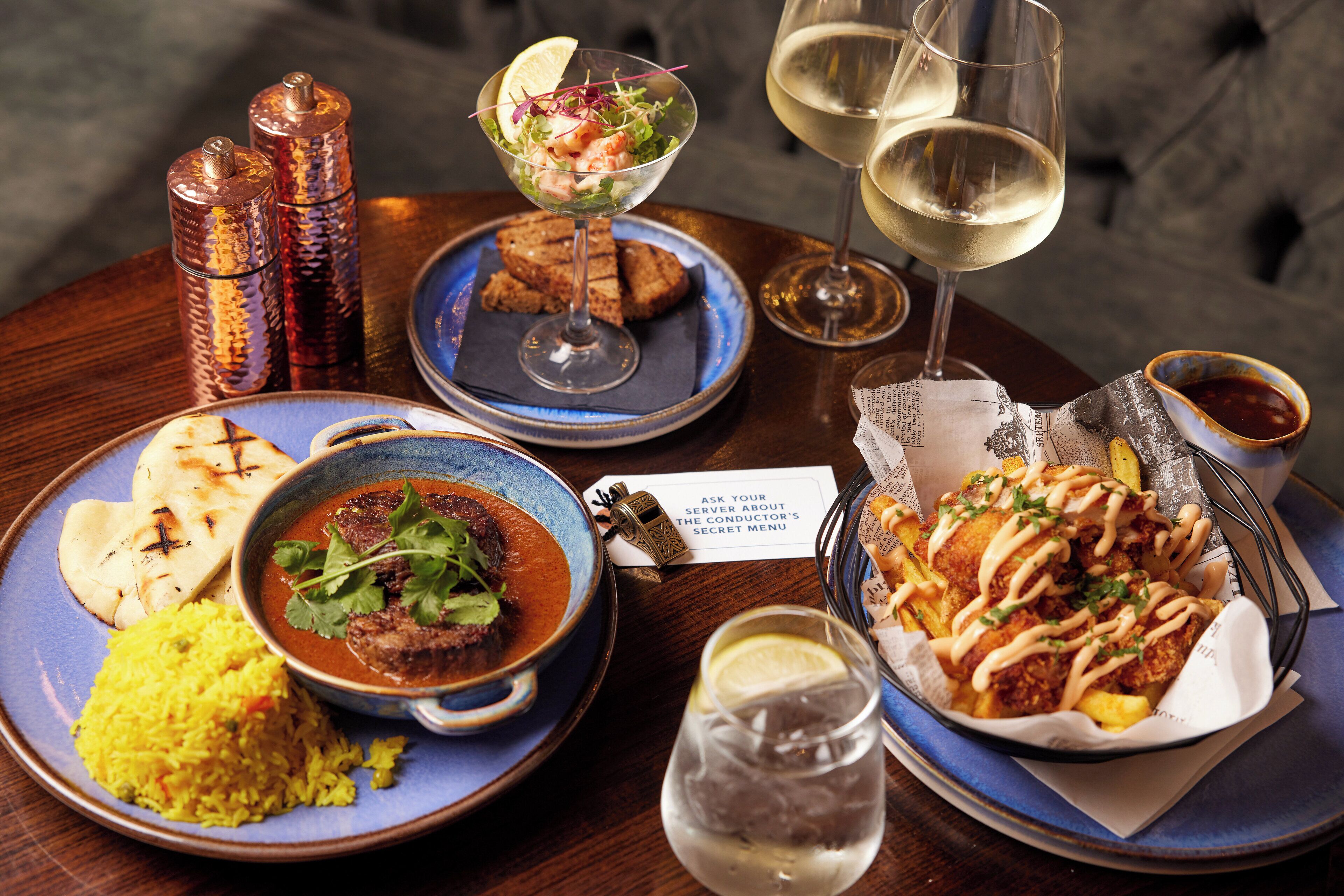 A table with various dishes, including a bowl of rice, a bowl of soup, grilled bread, and a basket of fries. There are also two glasses of white wine and a glass of water.