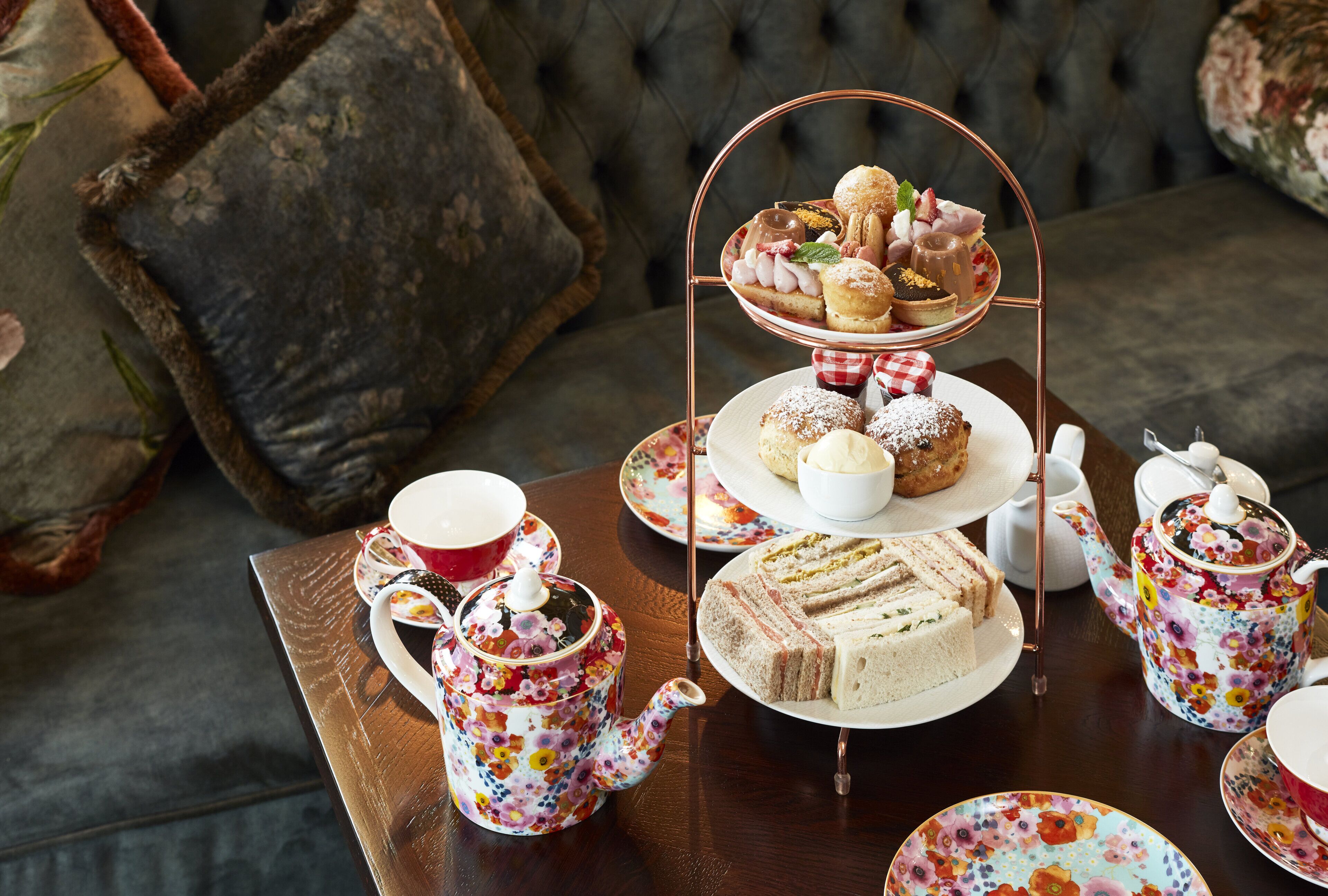 A tea set with floral patterns, a tiered dessert stand with pastries, sandwiches, and a teapot on a wooden table.