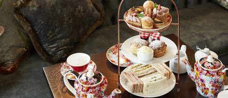 A tea set with floral patterns, a tiered dessert stand with pastries, sandwiches, and a teapot on a wooden table.
