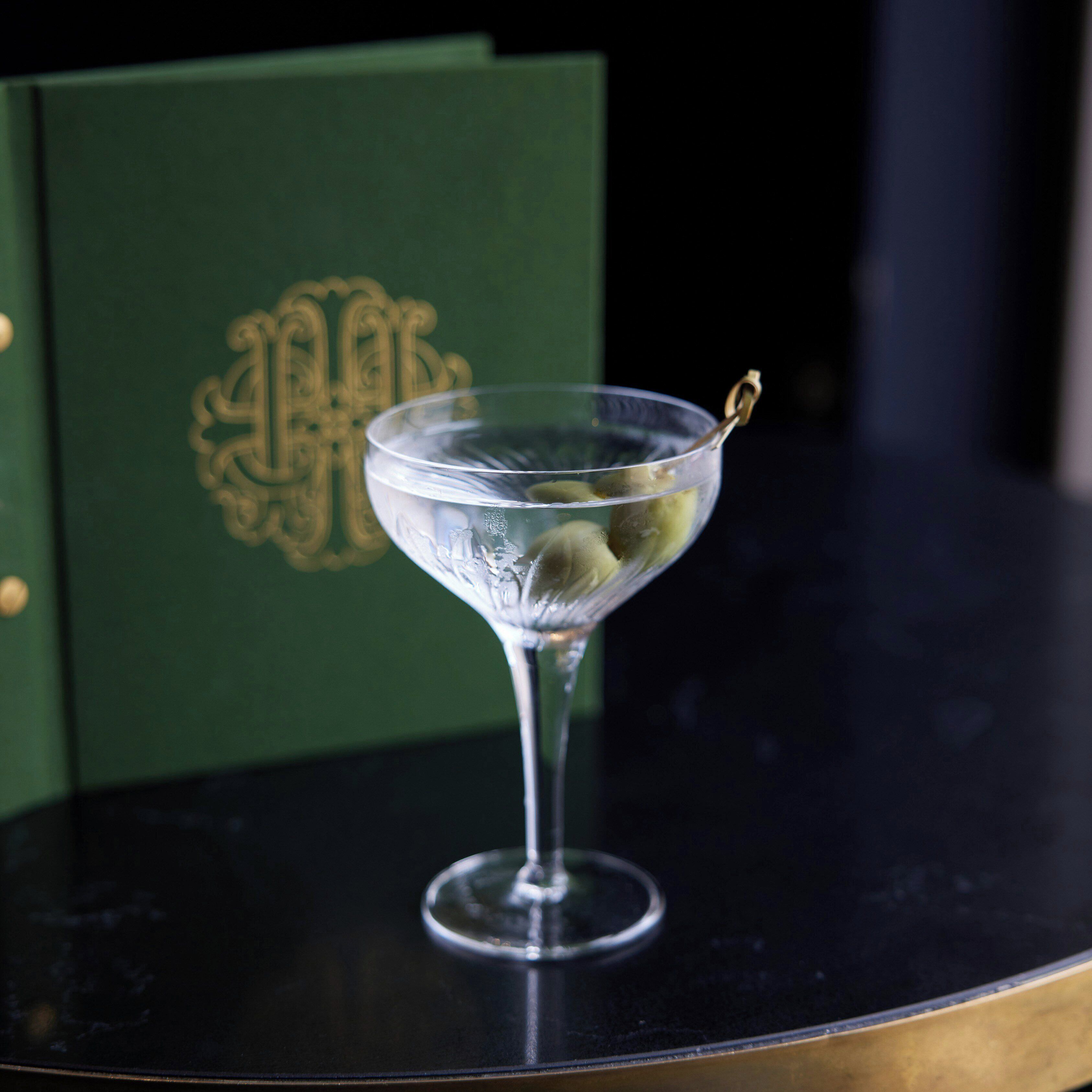 A martini glass with a garnish on a dark surface, possibly a table, with a green background featuring a gold emblem.