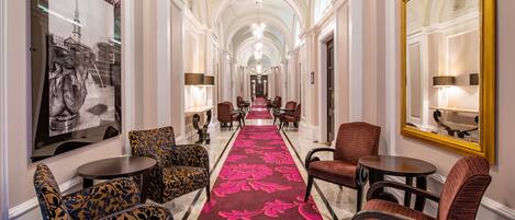 A hotel corridor with a red carpet, patterned chairs, and a chandelier.