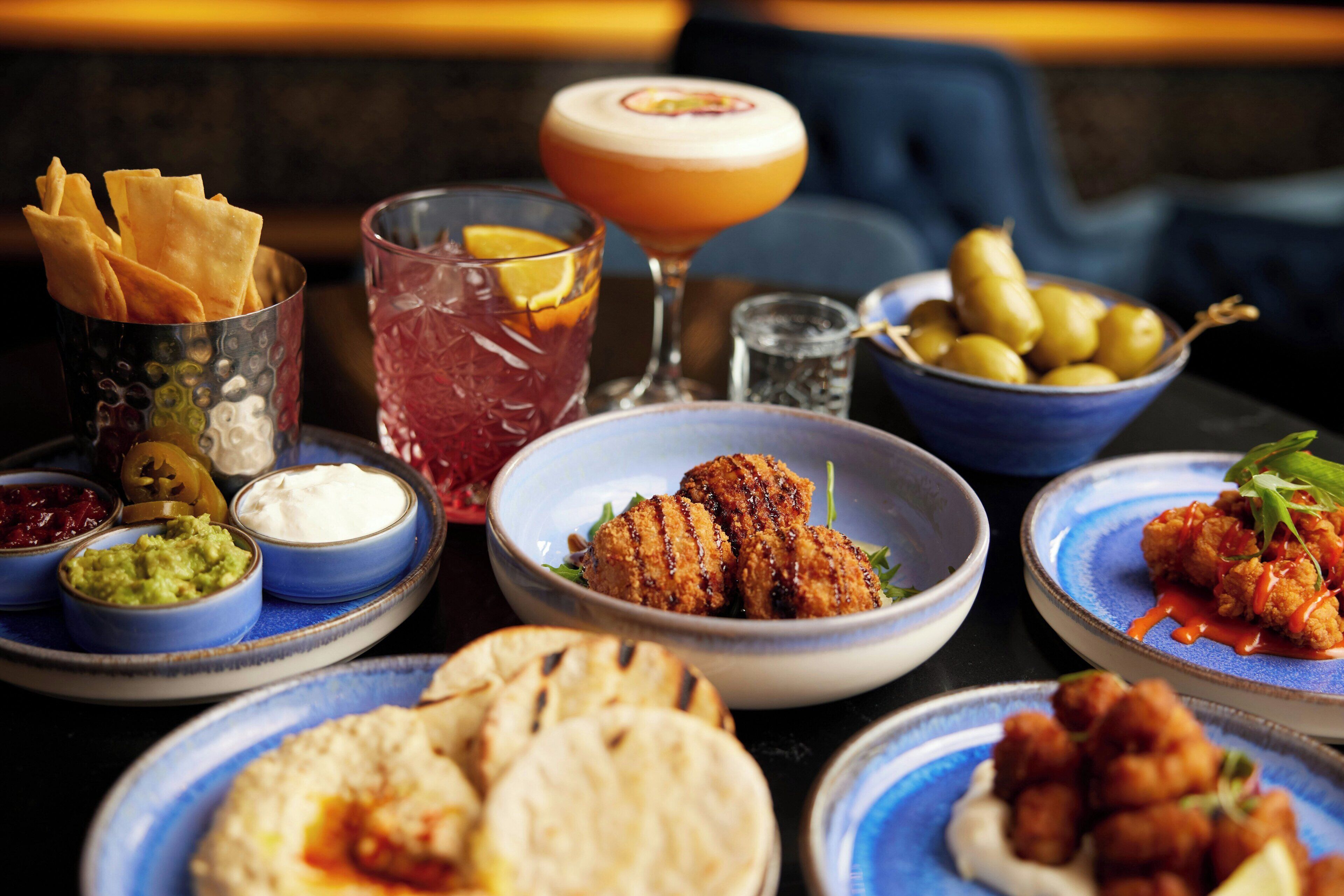 A variety of dishes and drinks on a table, including flatbreads, dips, and a cocktail.