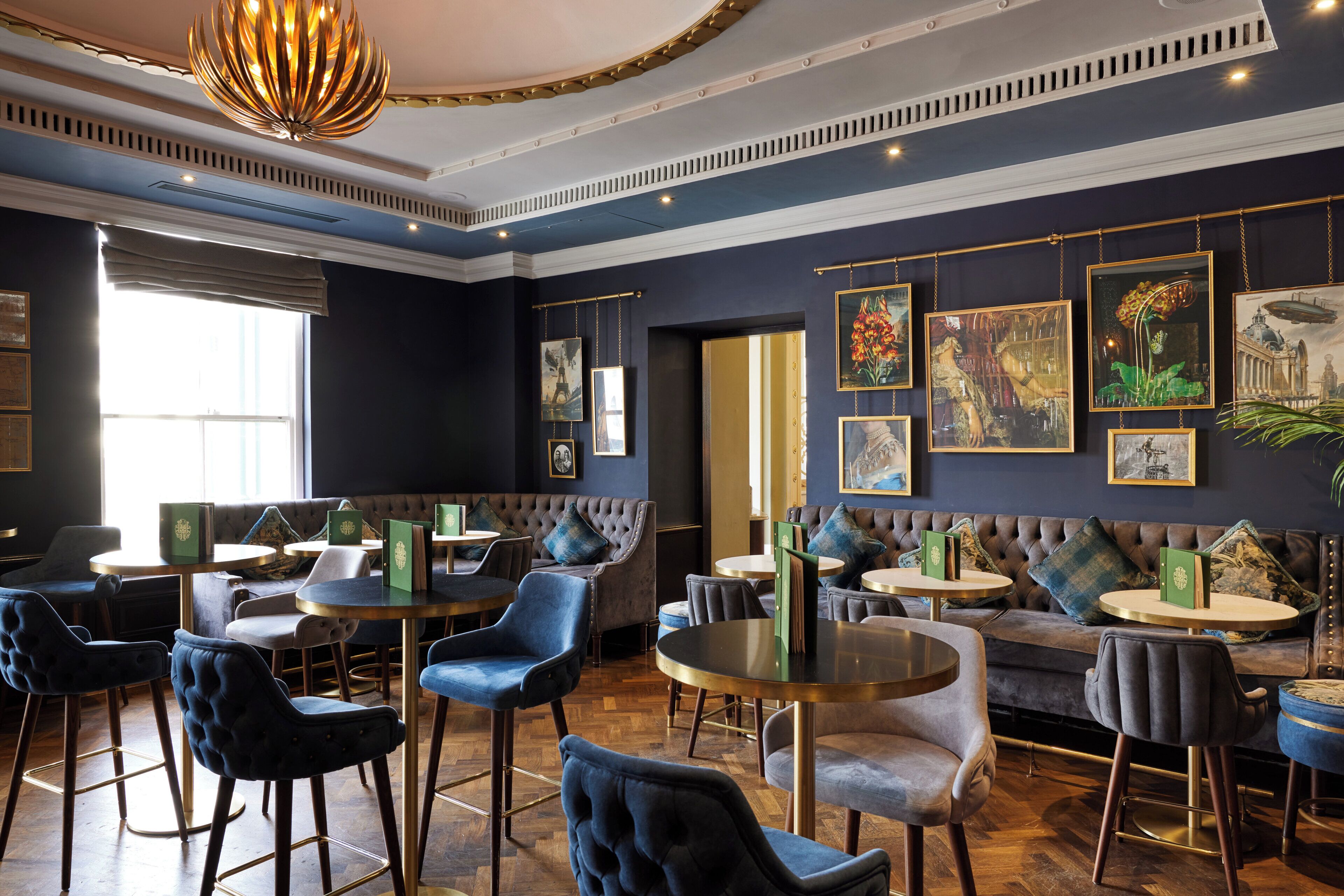A restaurant with round tables, blue upholstered chairs, and a dark blue wall with framed artwork.