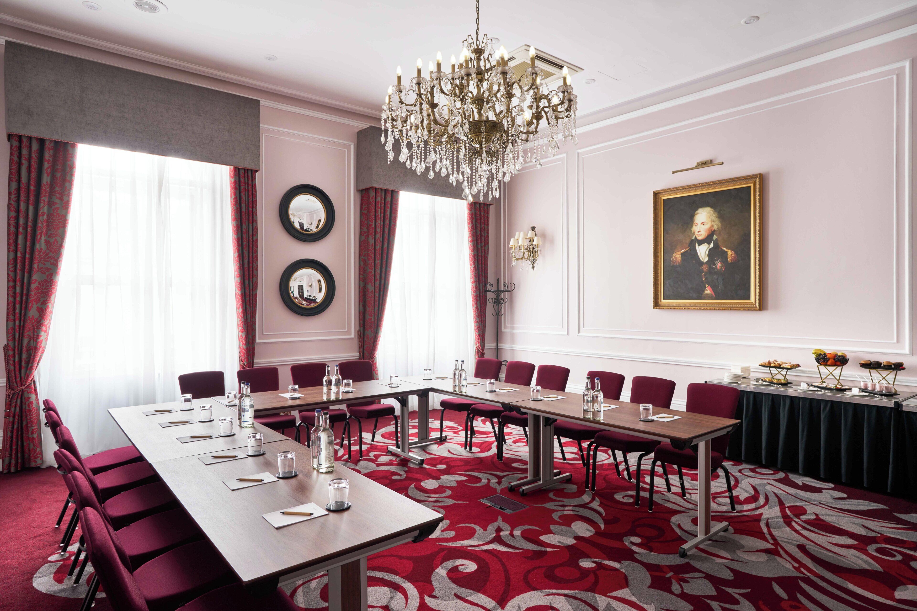 A conference room with a long table, red chairs, a chandelier, and a portrait on the wall.