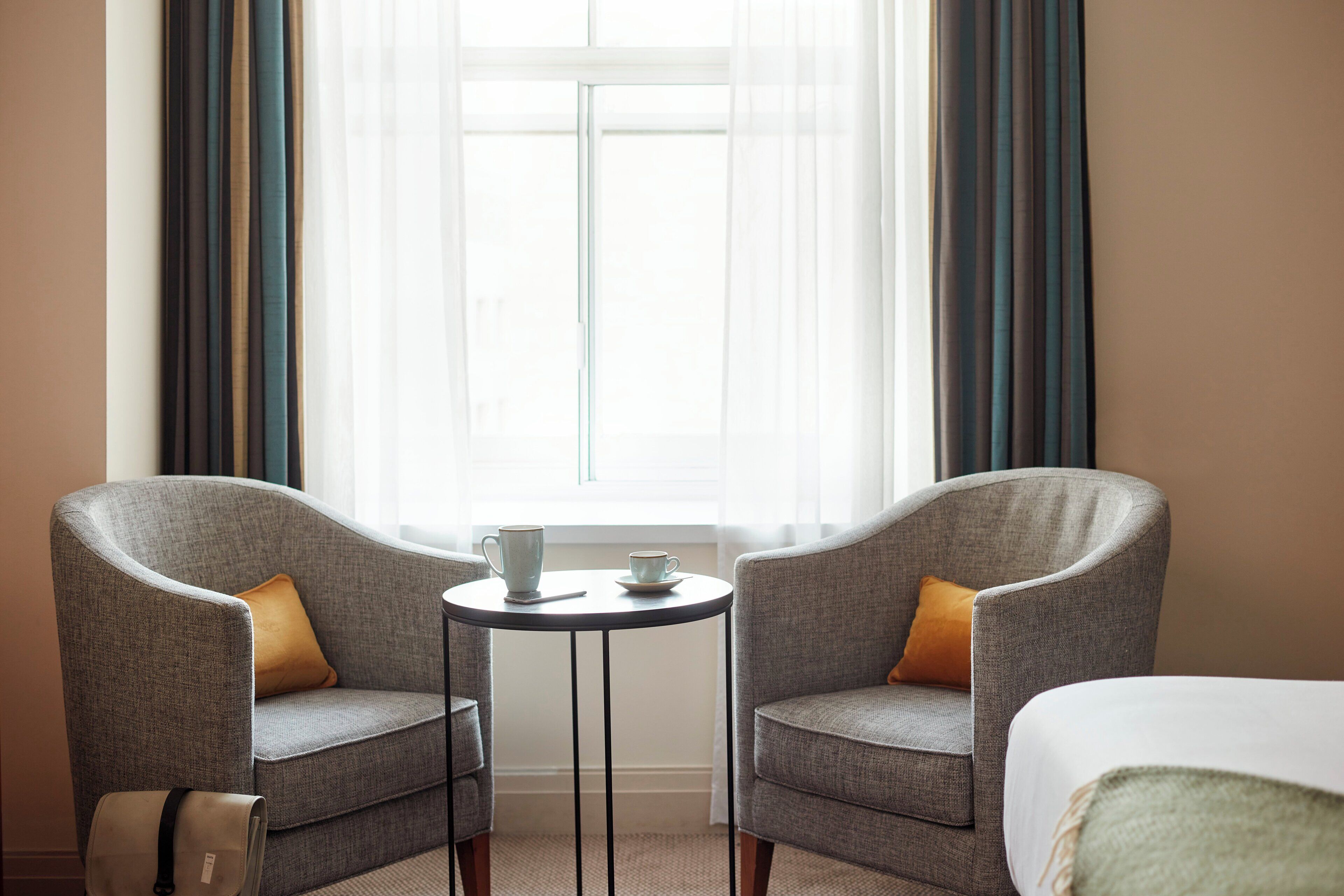 A hotel room with two armchairs, a small round table with a cup and saucer, and a bed with a green blanket.