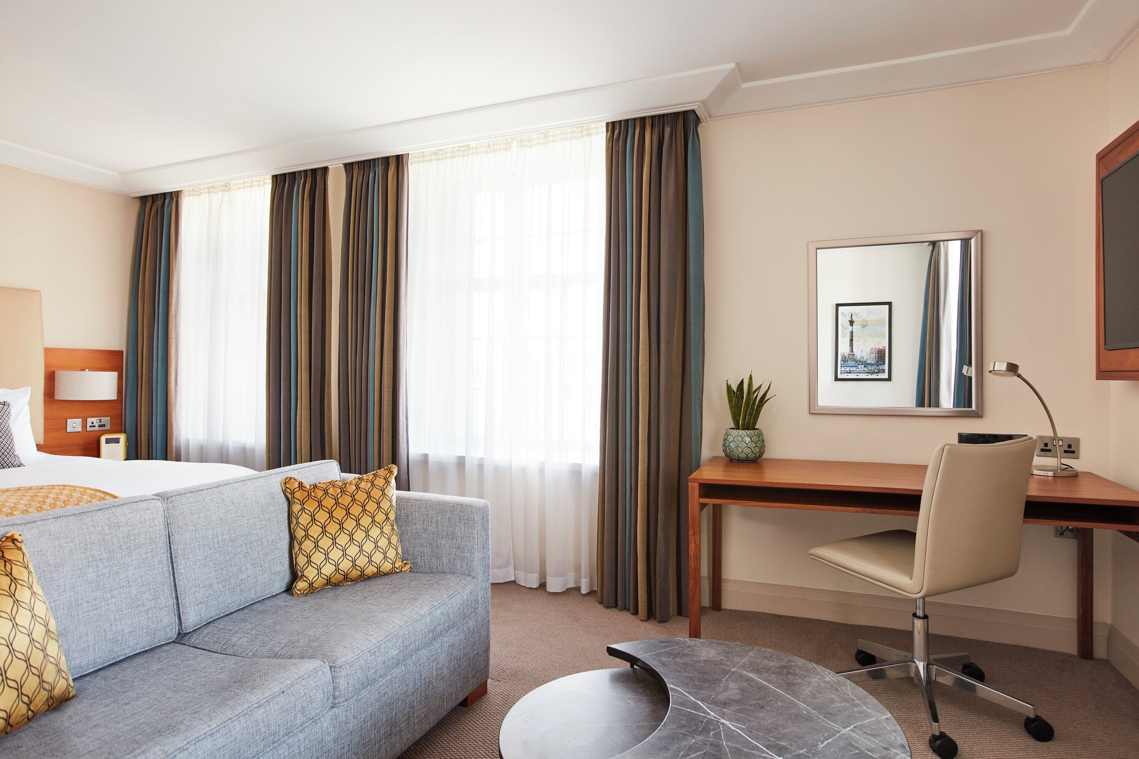 Une chambre d’hôtel avec un lit, un canapé gris agrémenté de coussins à motifs, un bureau avec une chaise, une petite table et un grand miroir au mur.