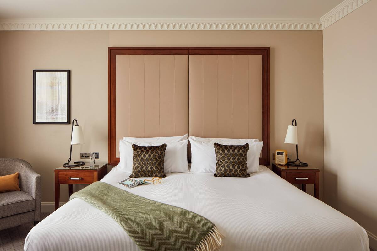 A hotel room with a large bed, two bedside tables, a chair, a framed picture on the wall, and a decorative ceiling medallion.