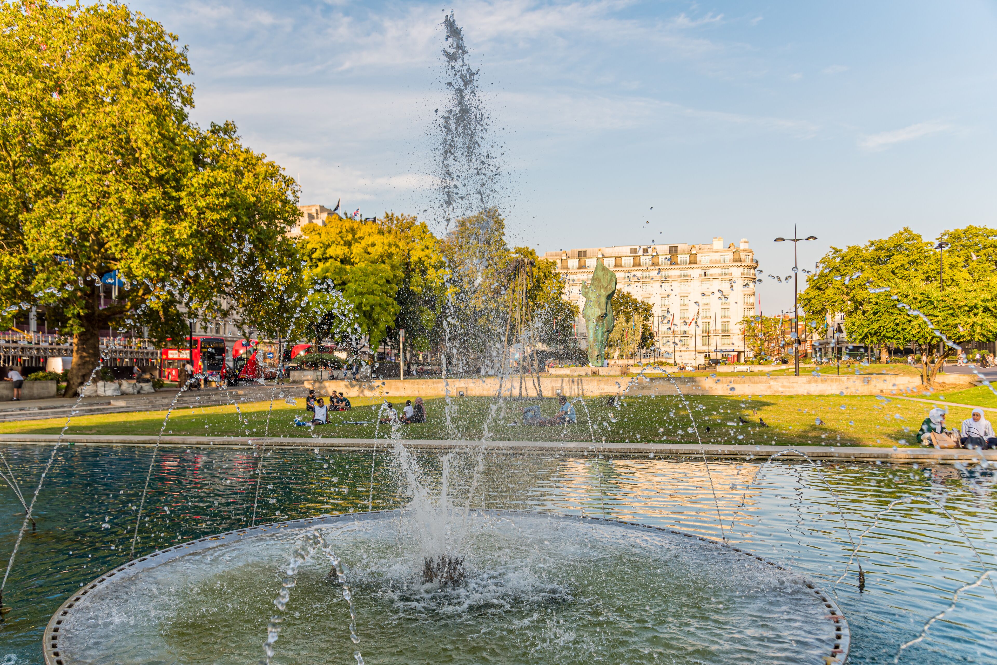 A fountain with water jets in a public park with trees and a building in the background.