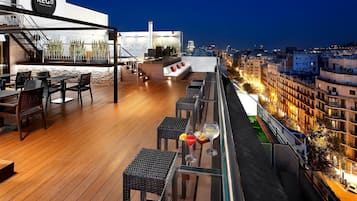 A rooftop terrace with wooden flooring, outdoor seating, and a city view at dusk.