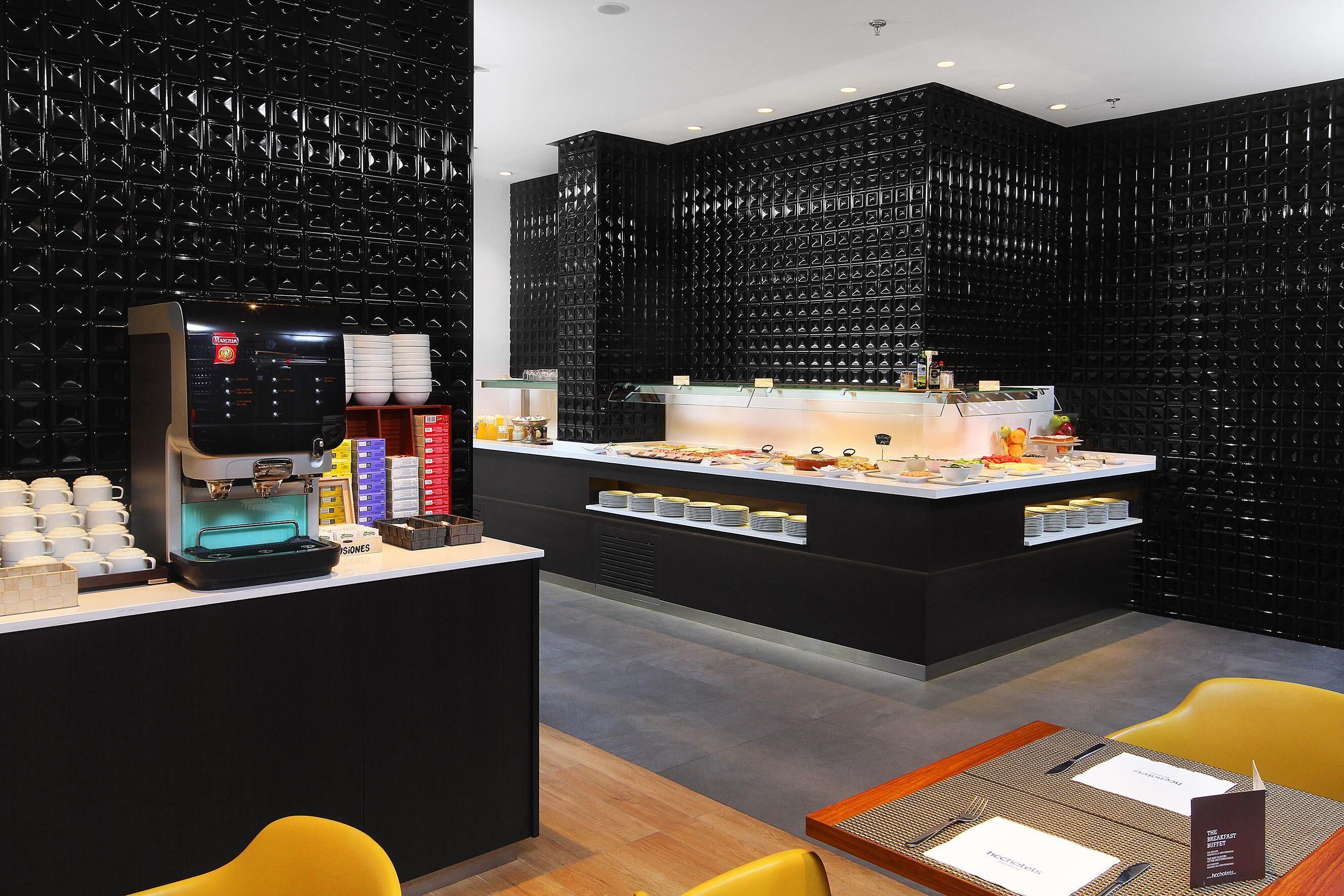 Un café moderno con una pared negra texturizada, una máquina de café y un mostrador de buffet con diversos alimentos.