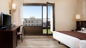 A hotel room with a bed, a desk with a TV, a chair, a balcony with a view of buildings, and a window with curtains. - Hotel HCC Regente (Barcelona)
