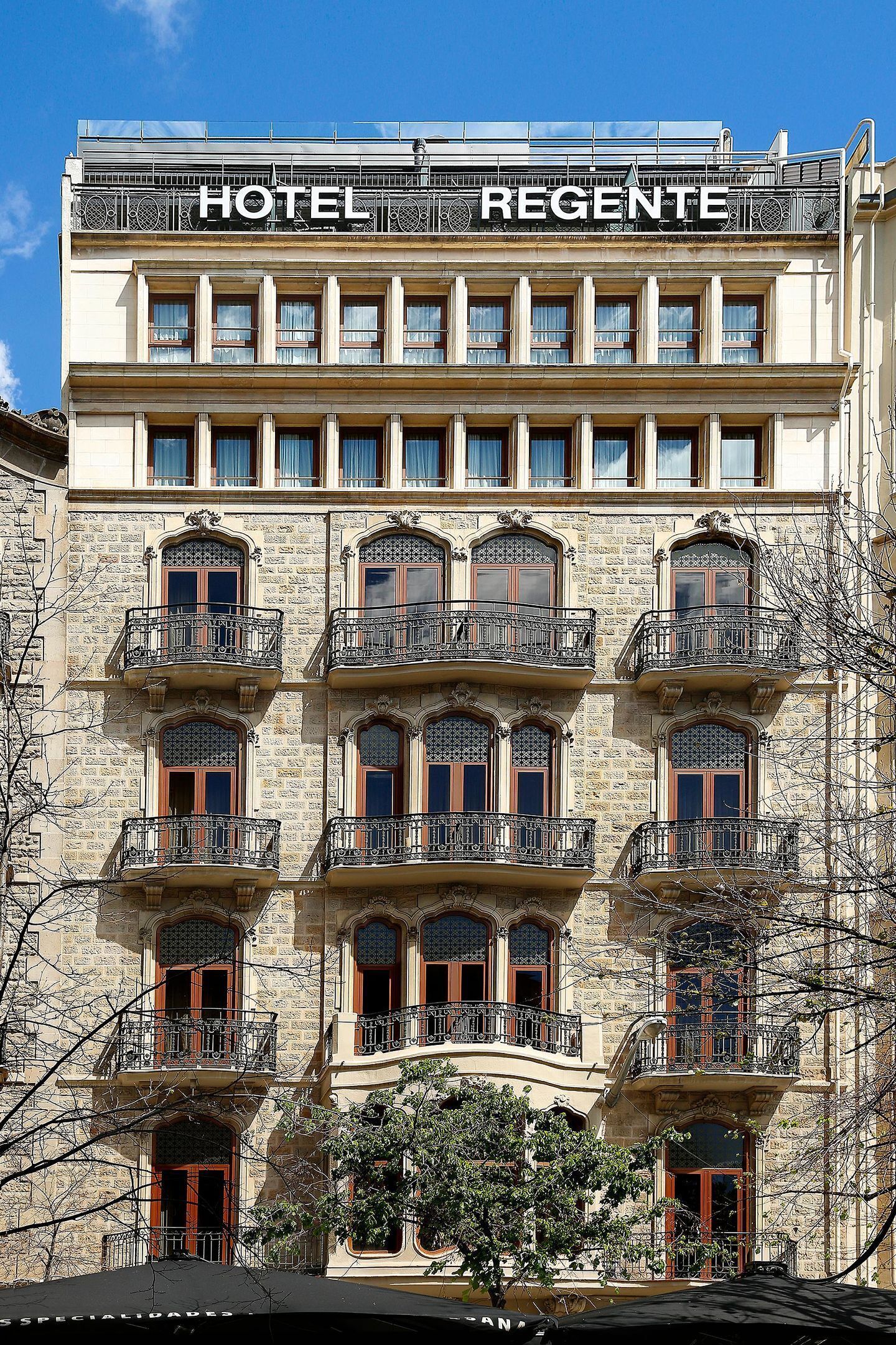 Un hotel a più piani con balconi, un cartello con la scritta "HOTEL REGENTE" e un cielo azzurro sullo sfondo.