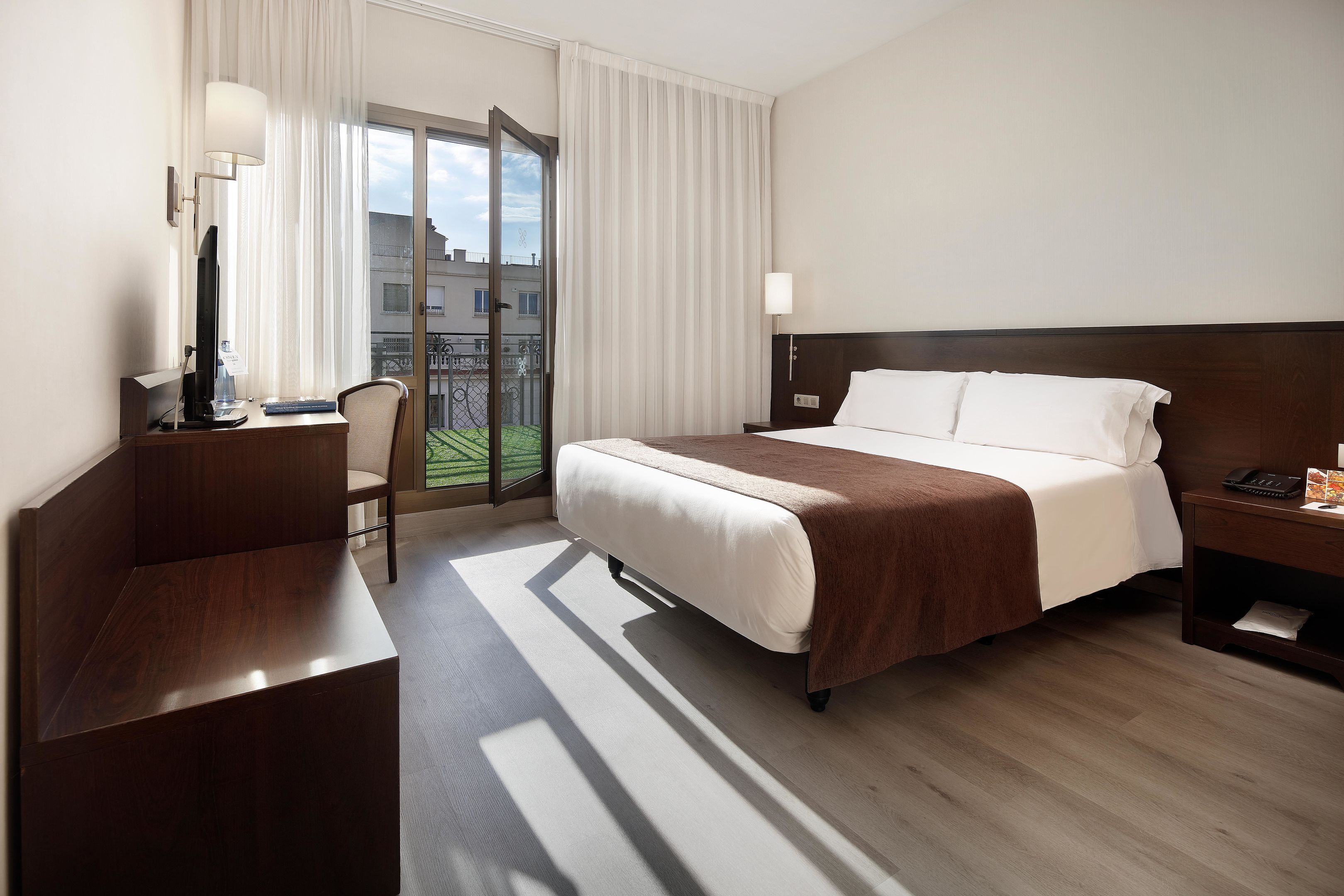 Habitación de hotel con cama, escritorio, silla, mesita de noche y vistas al exterior.