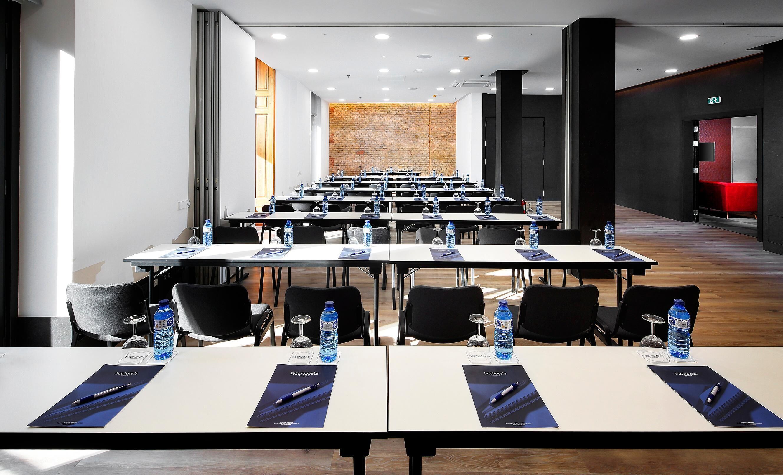 Un salón de conferencias con filas de mesas, sillas, botellas de agua y bolígrafos.