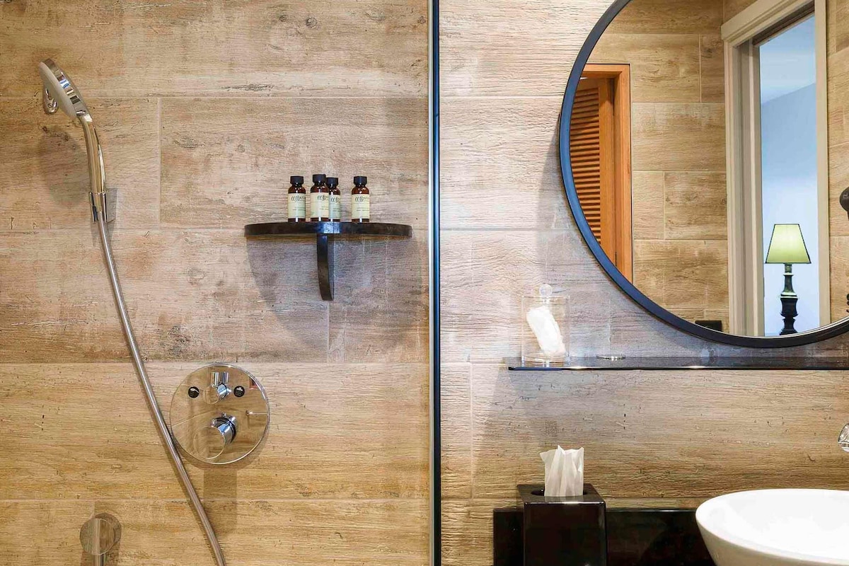 A bathroom with a wooden wall, a round mirror, a sink, and a shower with a handheld showerhead.