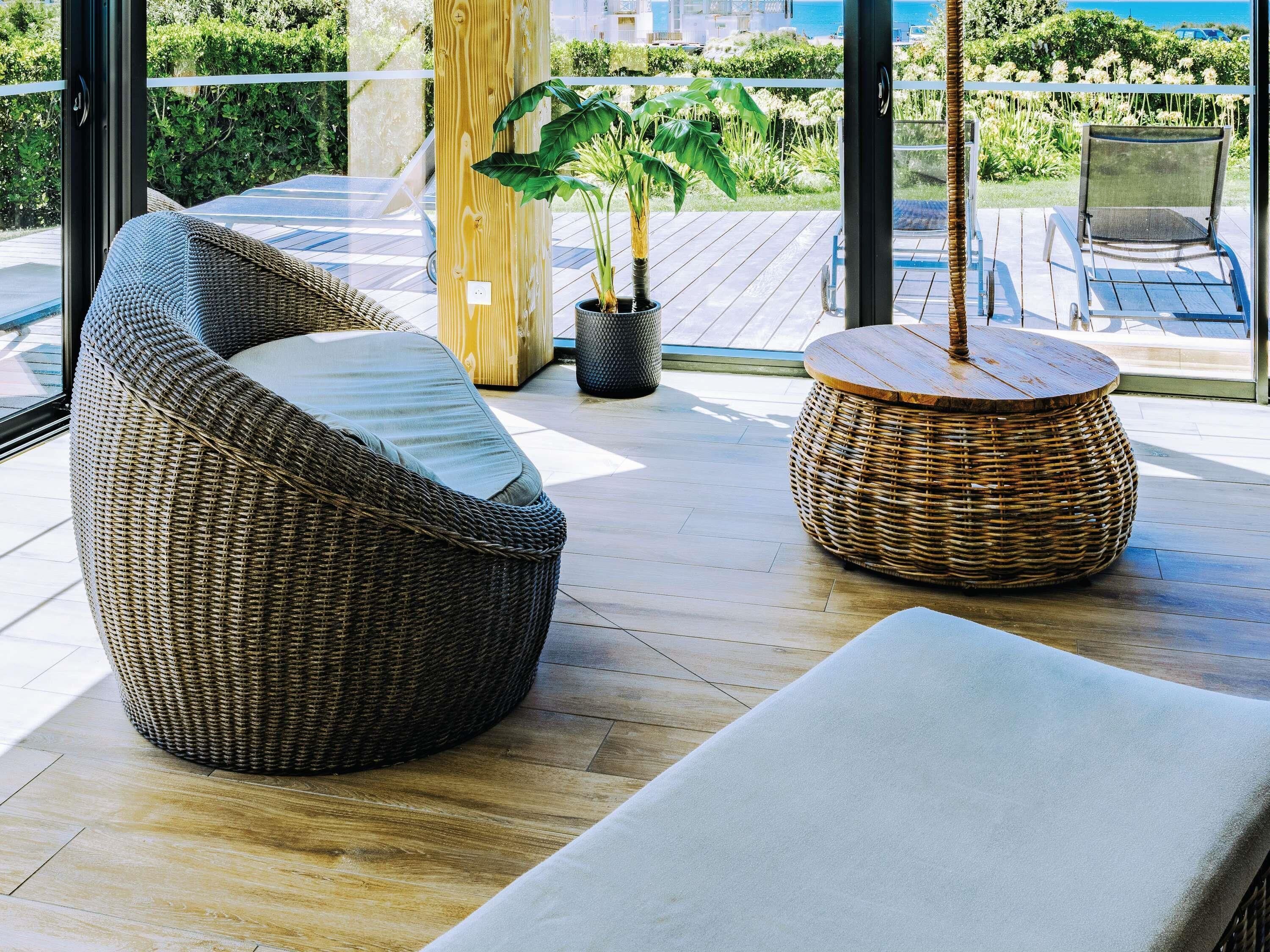 A wicker sofa, a round wicker table, and a potted plant on a wooden deck with a view of the sea.