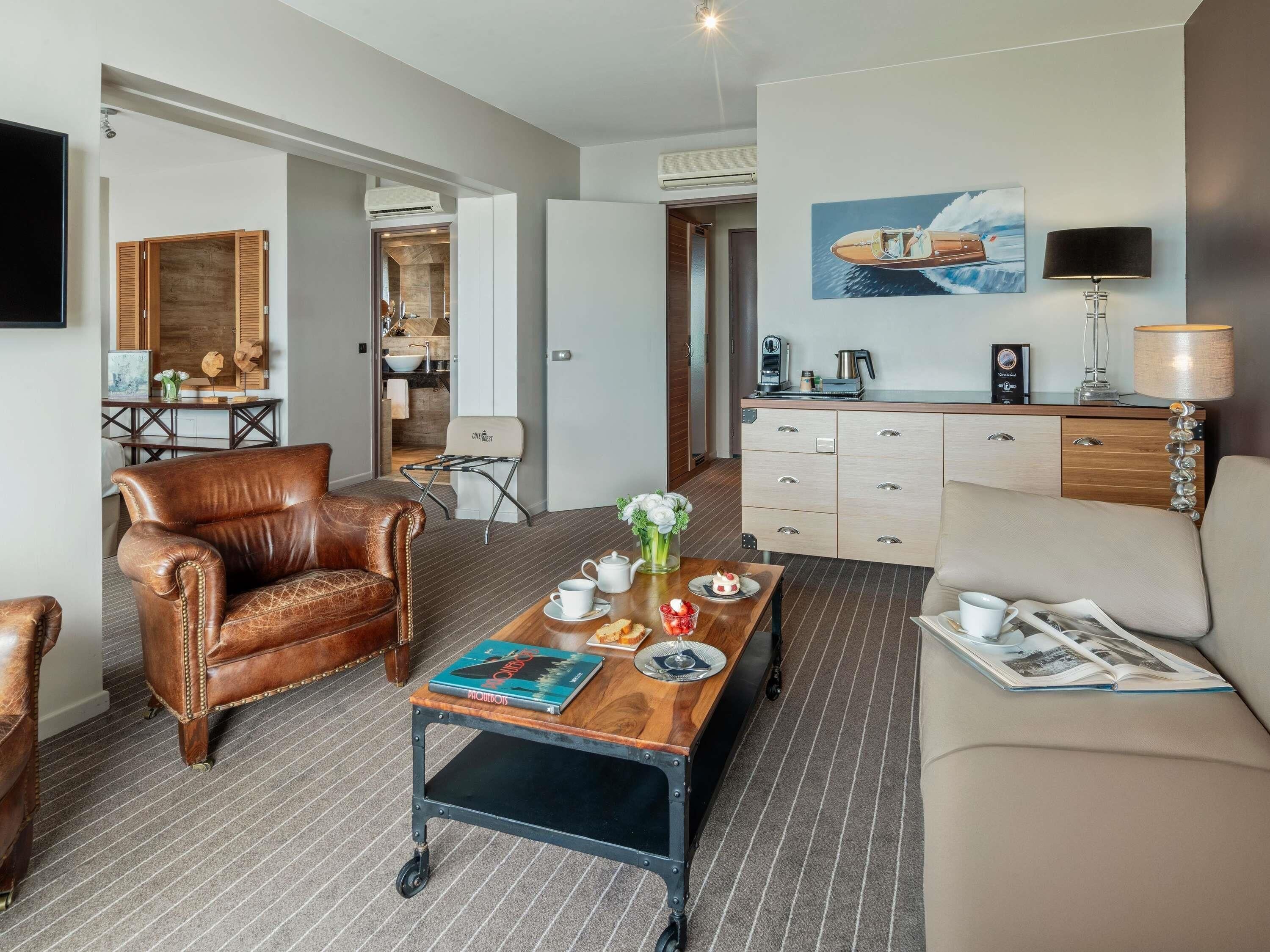 A modern hotel room with a leather armchair, a sofa, a coffee table with books, a dining table, and a kitchenette.