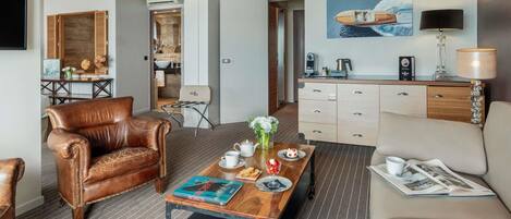 A modern hotel room with a leather armchair, a sofa, a coffee table with books, a dining table, and a kitchenette.