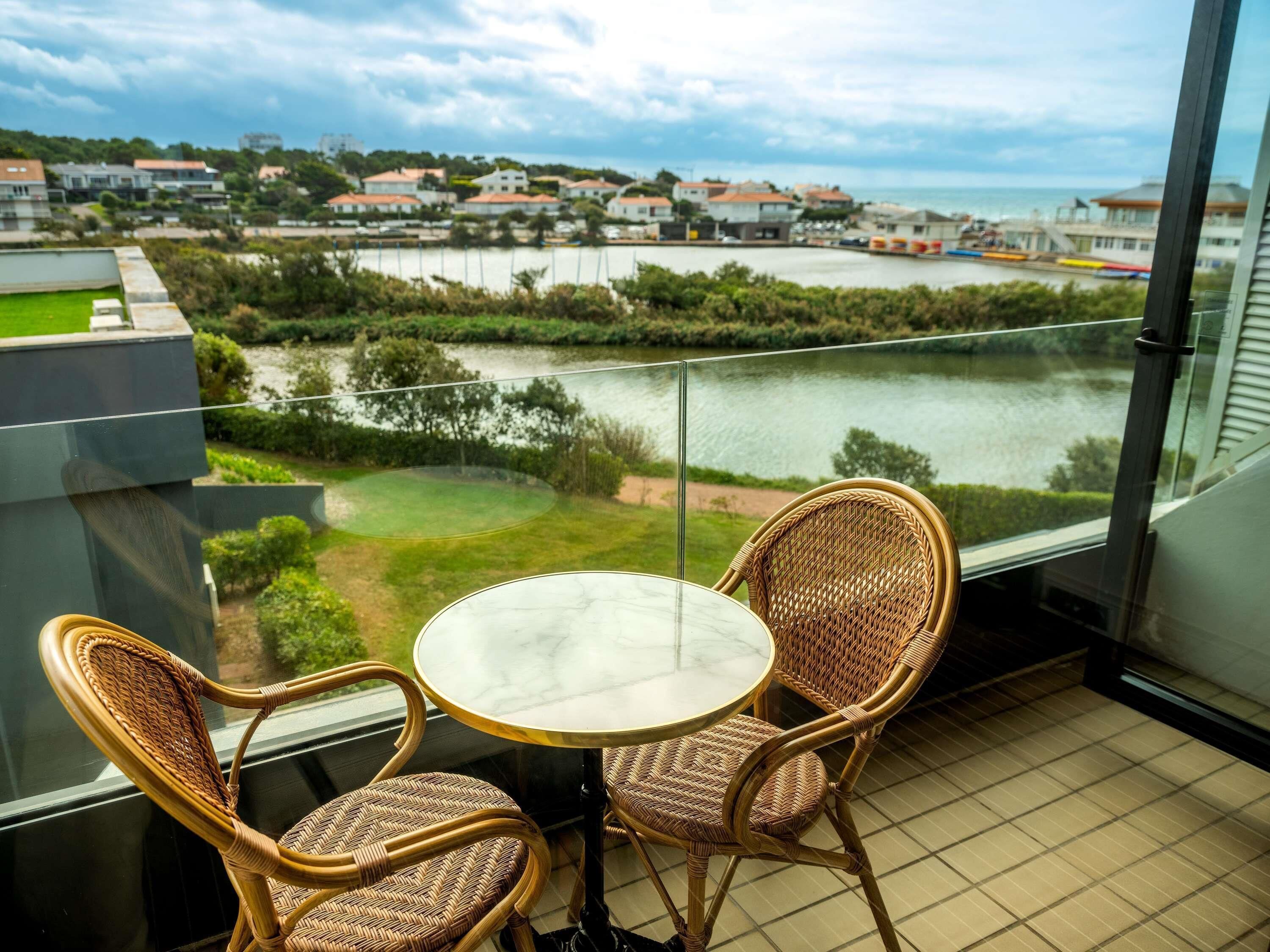 Un balcon avec des fauteuils en osier et une petite table, donnant sur une étendue d’eau et une zone résidentielle.