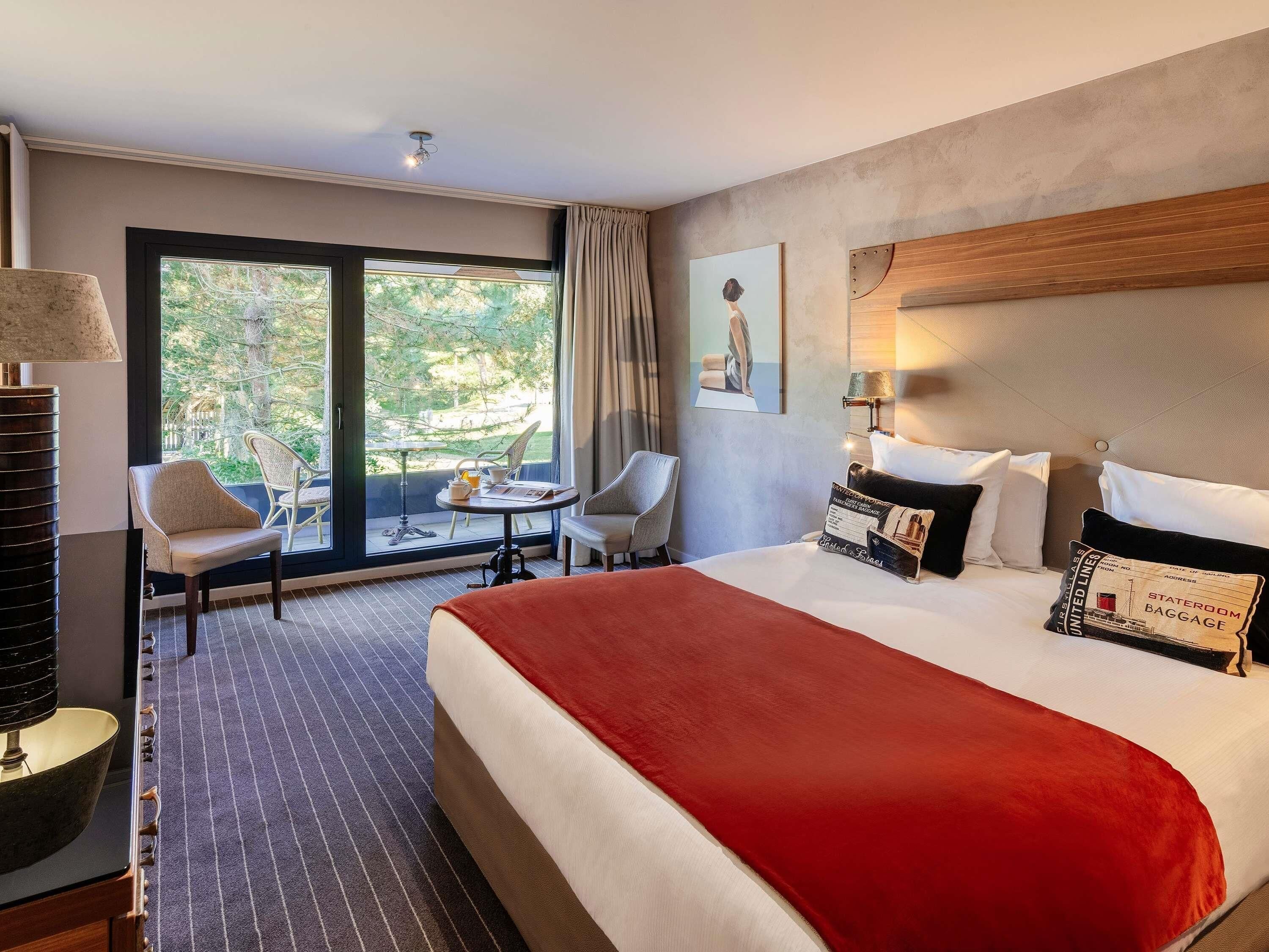 A hotel room with a large bed, two chairs, a table, and a view of trees through the window.