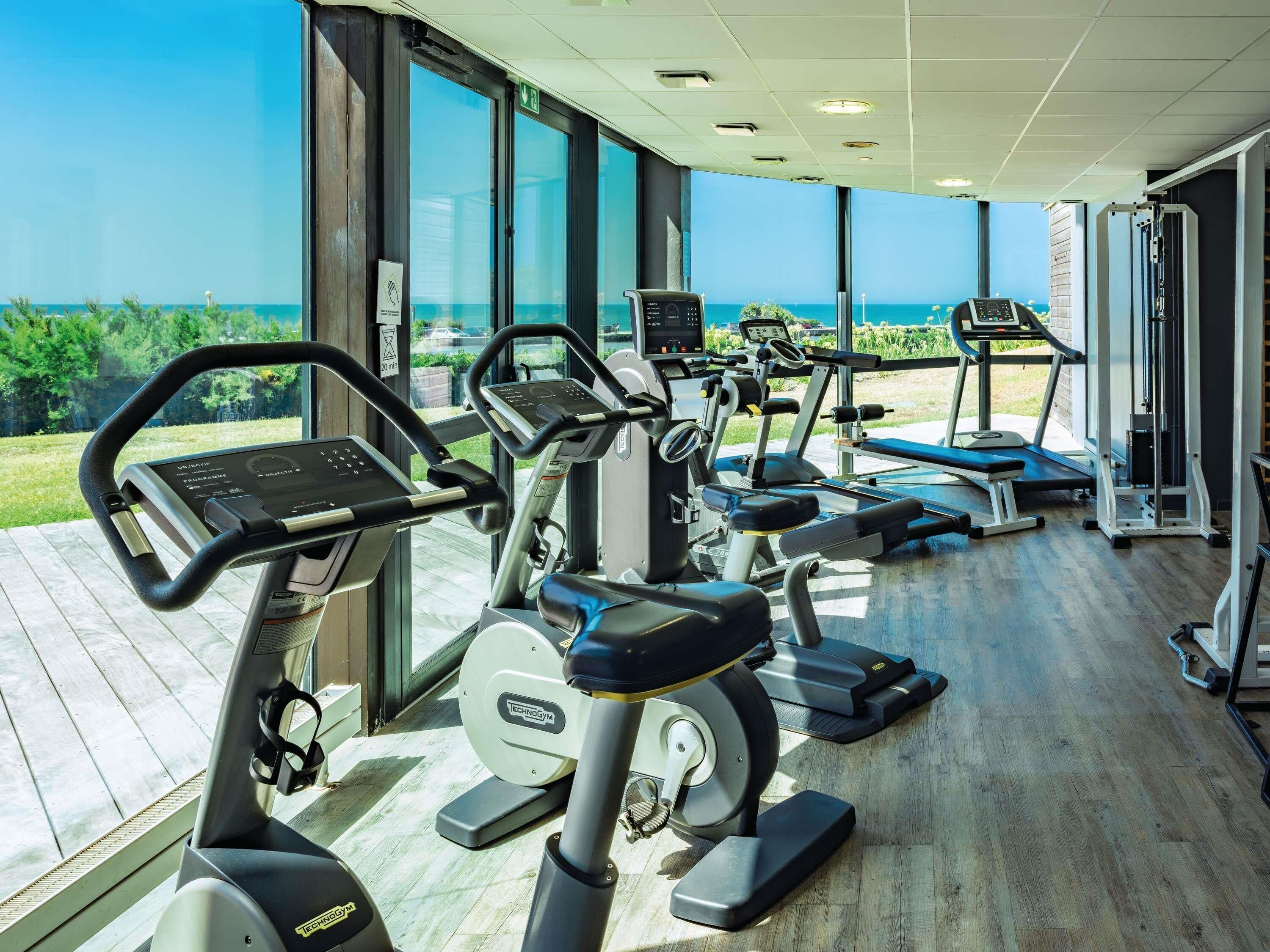 Ett gym med konditionsmaskiner, en bänk och en styrketräningsmaskin, med utsikt över havet genom stora fönster.