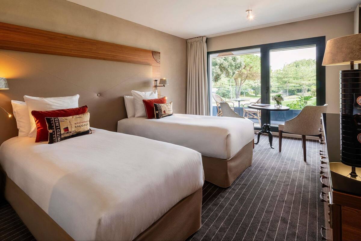Une chambre d’hôtel avec deux lits, un bureau, une chaise et une vue sur l’extérieur.