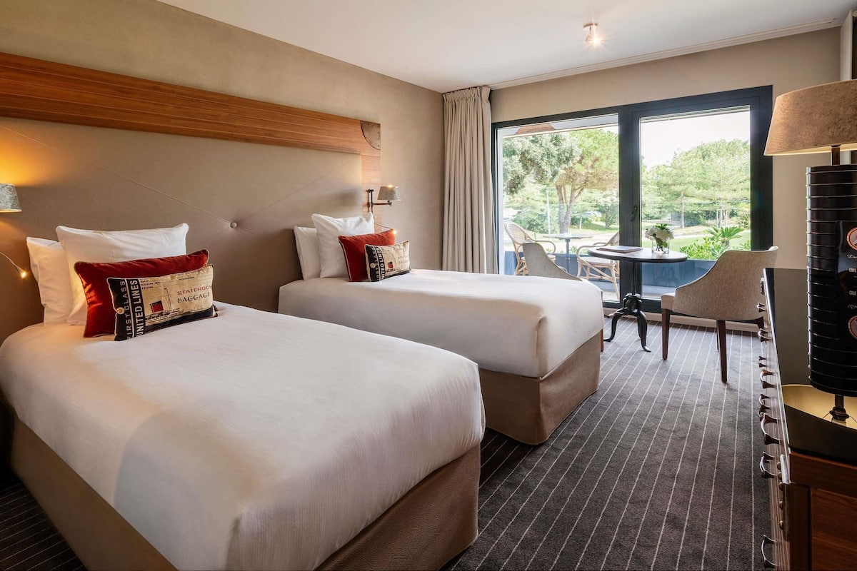 A hotel room with two beds, a desk, a chair, and a view of the outdoors.