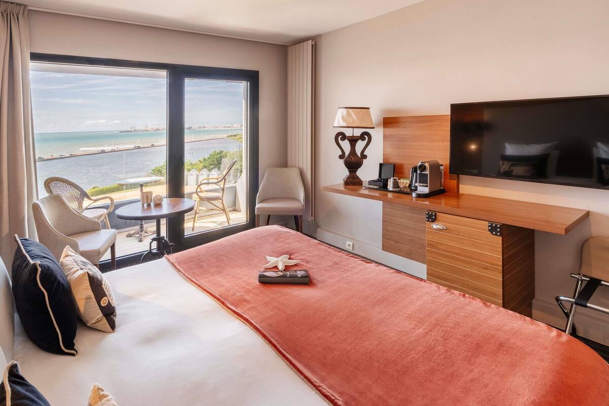 Une chambre d’hôtel avec un lit, un bureau, une chaise, une télévision et une vue sur la mer.