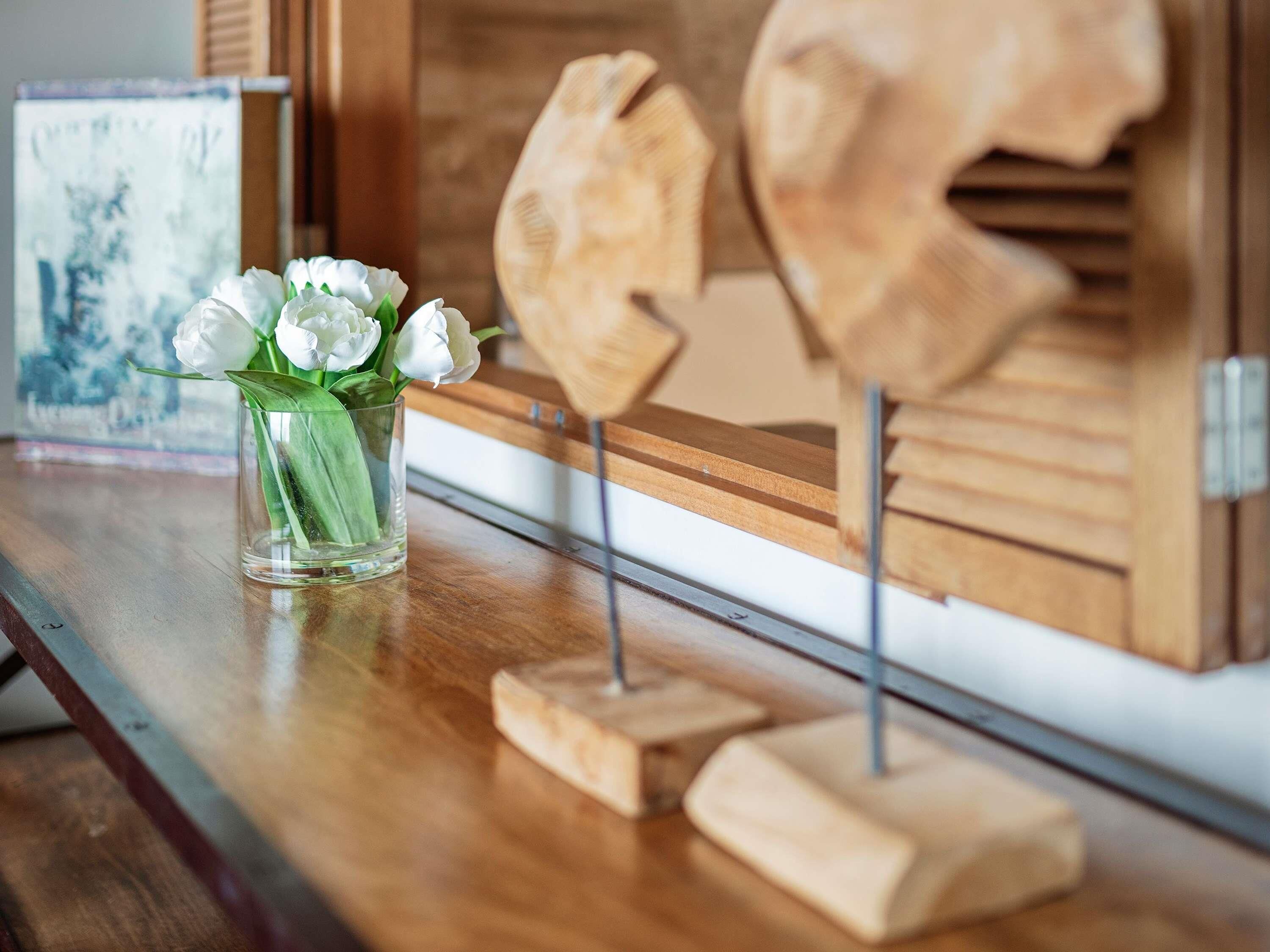 A wooden table with a clear glass vase containing white flowers, a framed picture, and wooden head sculptures.