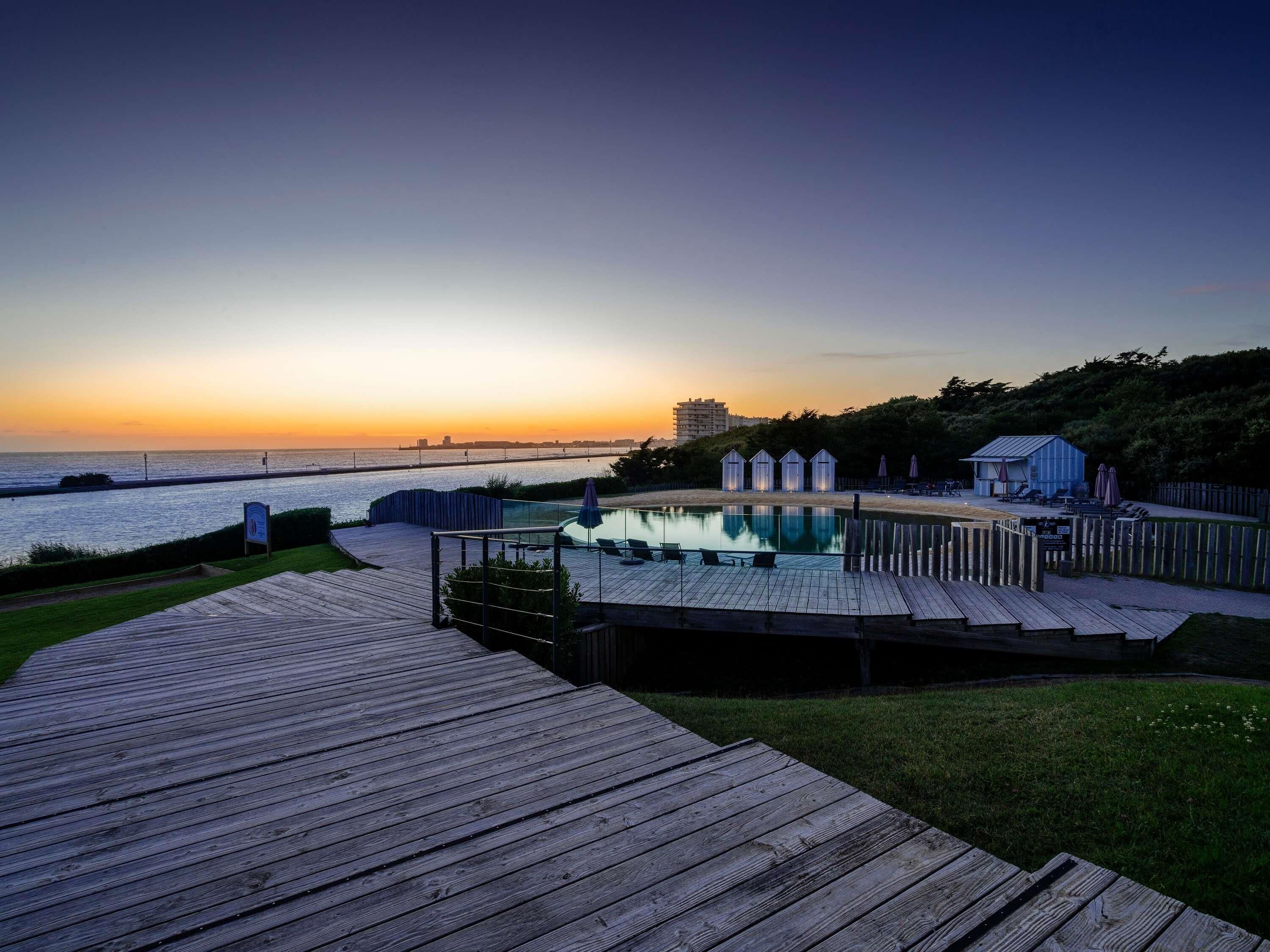 A wooden deck leading to a pool with a view of the sunset over the water.