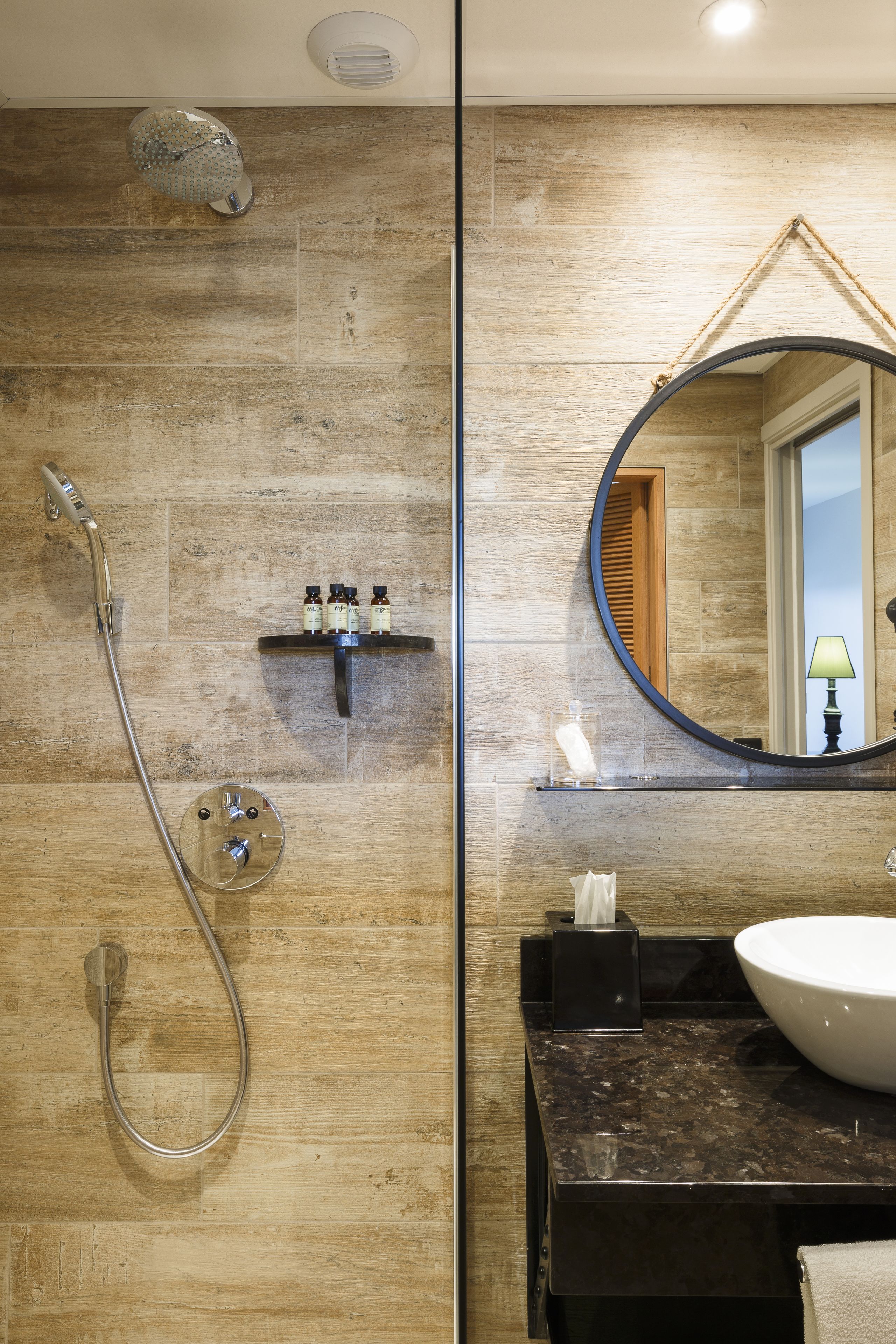 Une salle de bain avec un mur en bois, un miroir rond, un lavabo et une douche équipée d’un pommeau de douche à main.