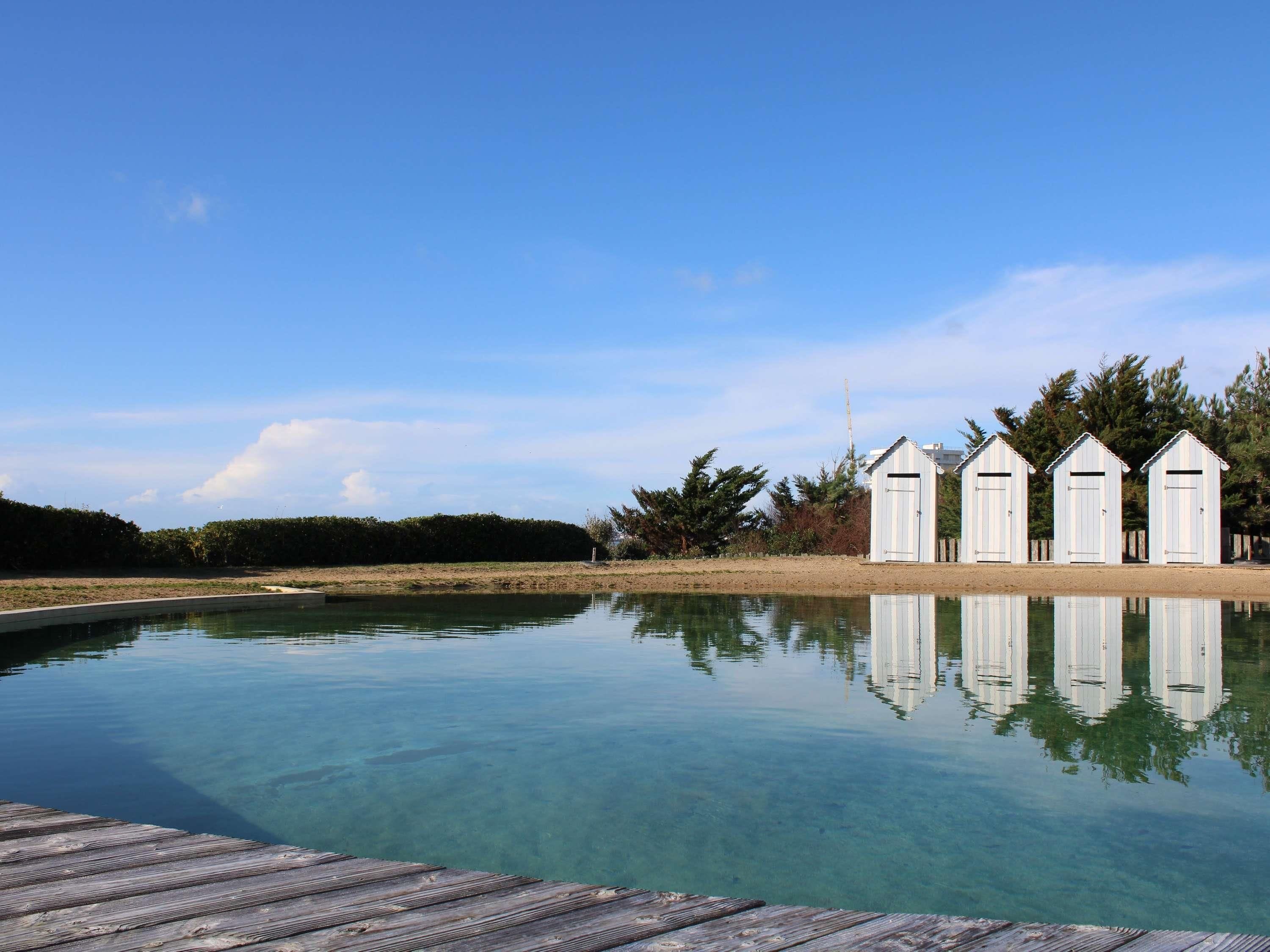 A clear pool with a wooden deck in the foreground, surrounded by a sandy beach and a row of white beach huts.
