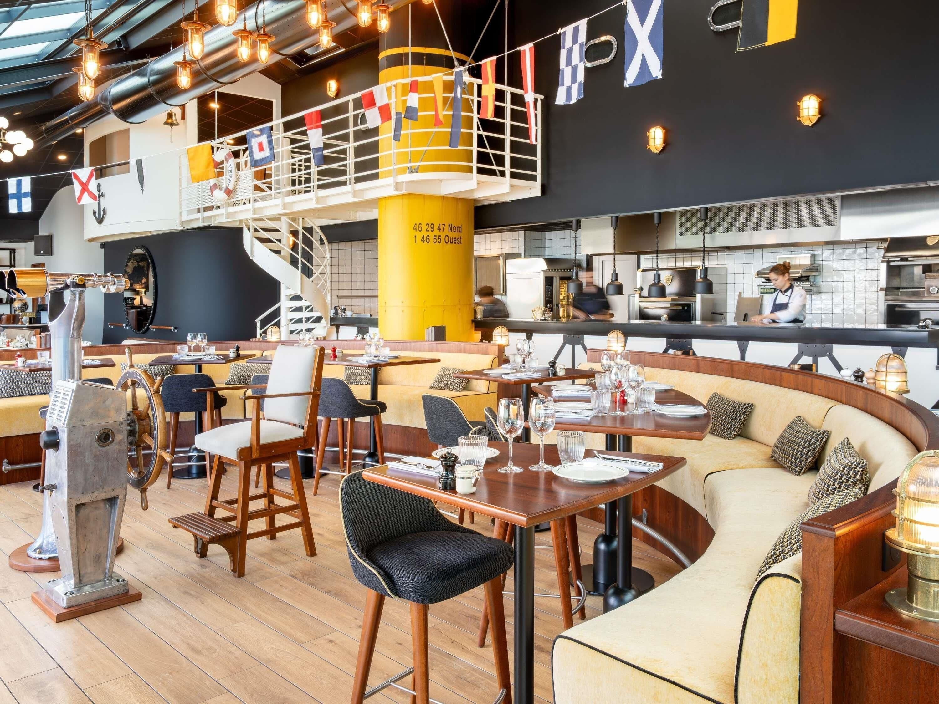 Un restaurant avec un plafond haut, des conduits apparents et un mélange de types de sièges, notamment des tables et un canapé incurvé. On aperçoit un bar à l’arrière-plan.
