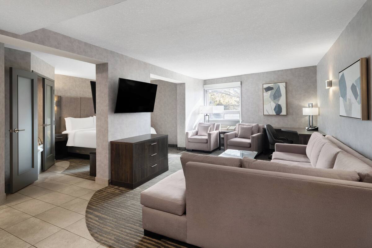 A modern hotel room with a large bed, a sofa, two armchairs, a flat-screen TV mounted on the wall, and a desk with a lamp.