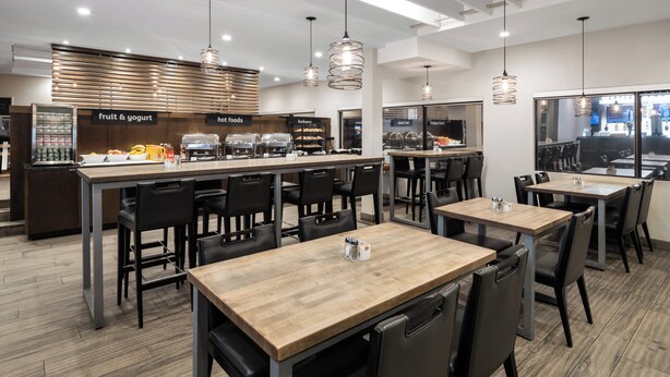 A modern cafe with a central counter offering fruit, yogurt, and hot foods. There are multiple seating areas with tables and chairs.