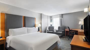 A hotel room with a large bed, a desk, two armchairs, a television, and a lamp.