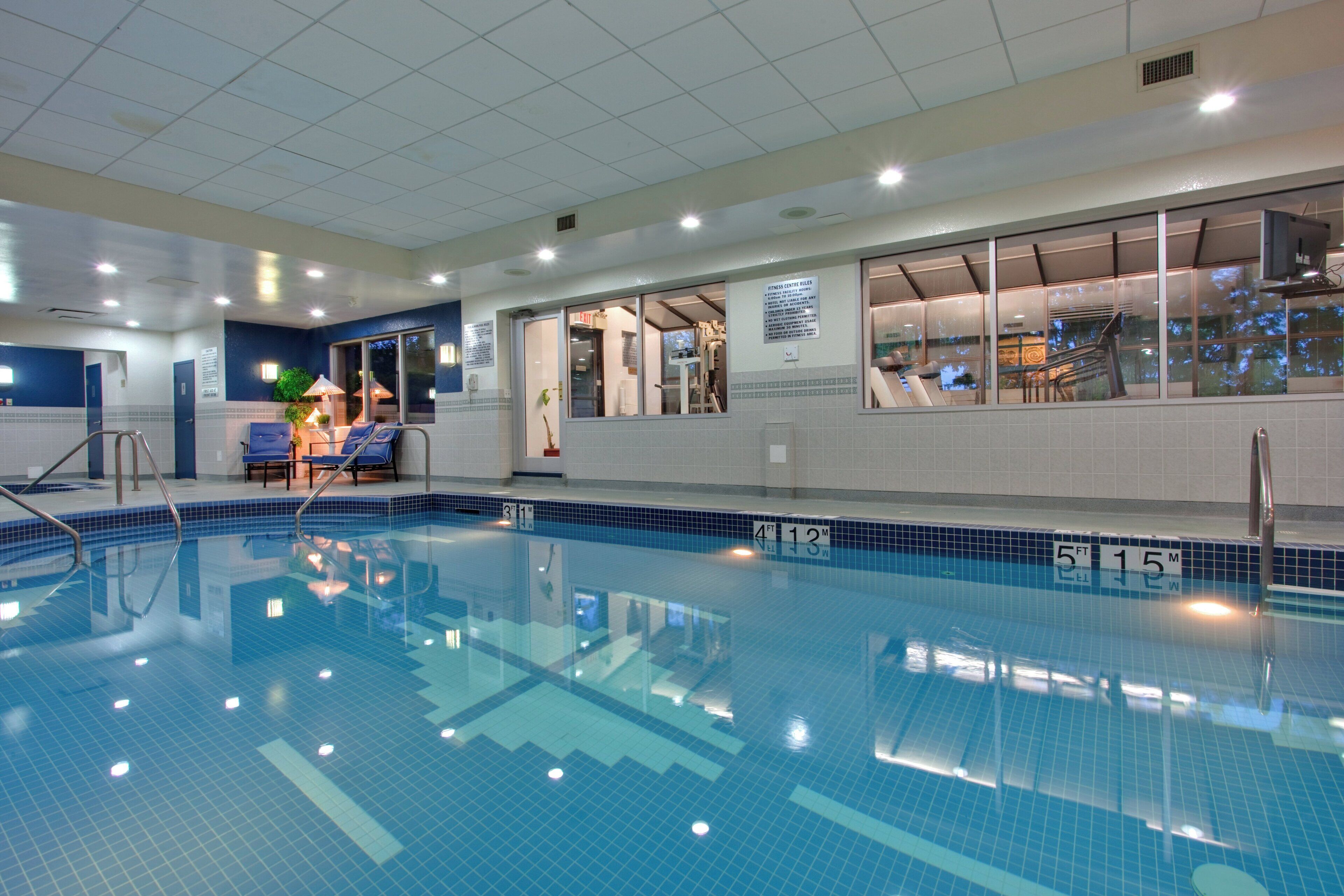 An indoor swimming pool with clear water, marked depth lines, and a sunlit area with lounge chairs.