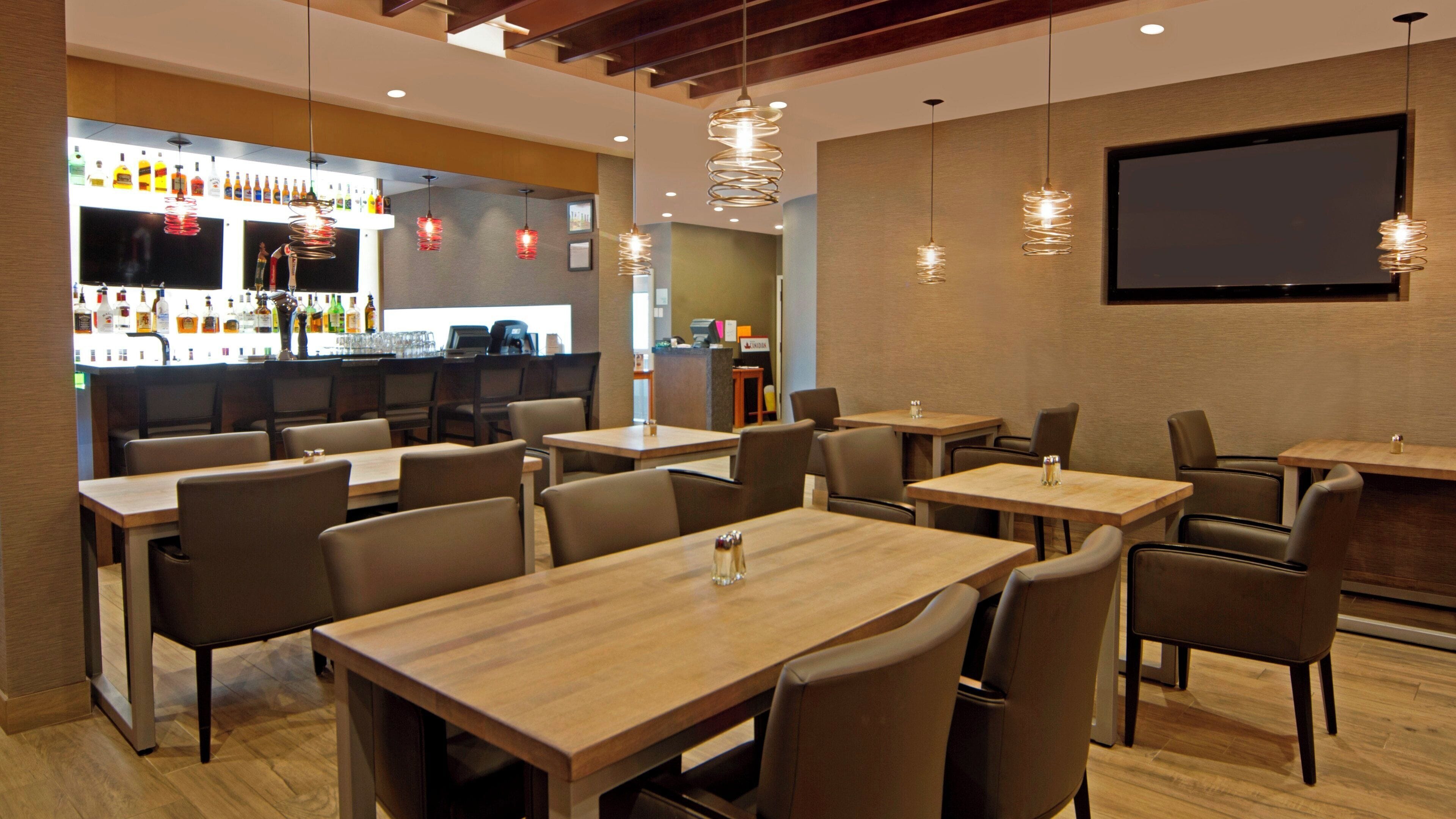 A restaurant with multiple tables and chairs, a bar area with various bottles, and hanging pendant lights.