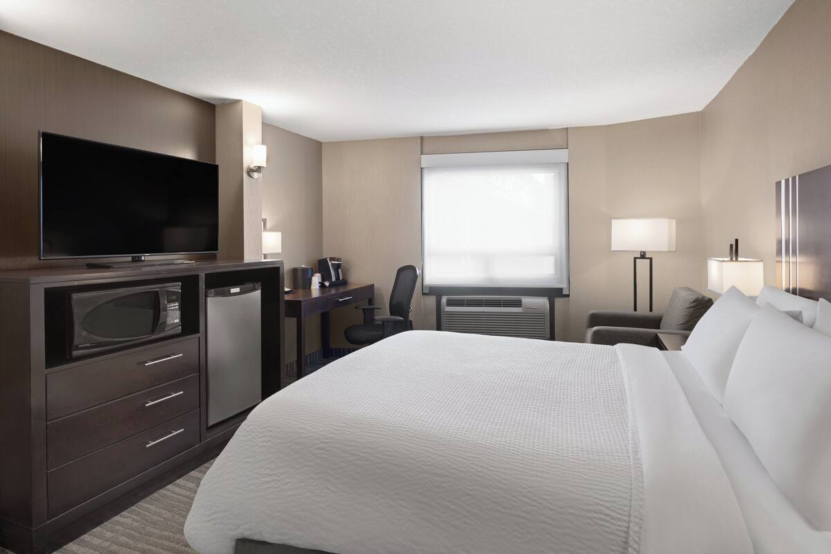 A hotel room with a bed, a desk with a computer, a TV, a microwave, a coffee maker, and a window.