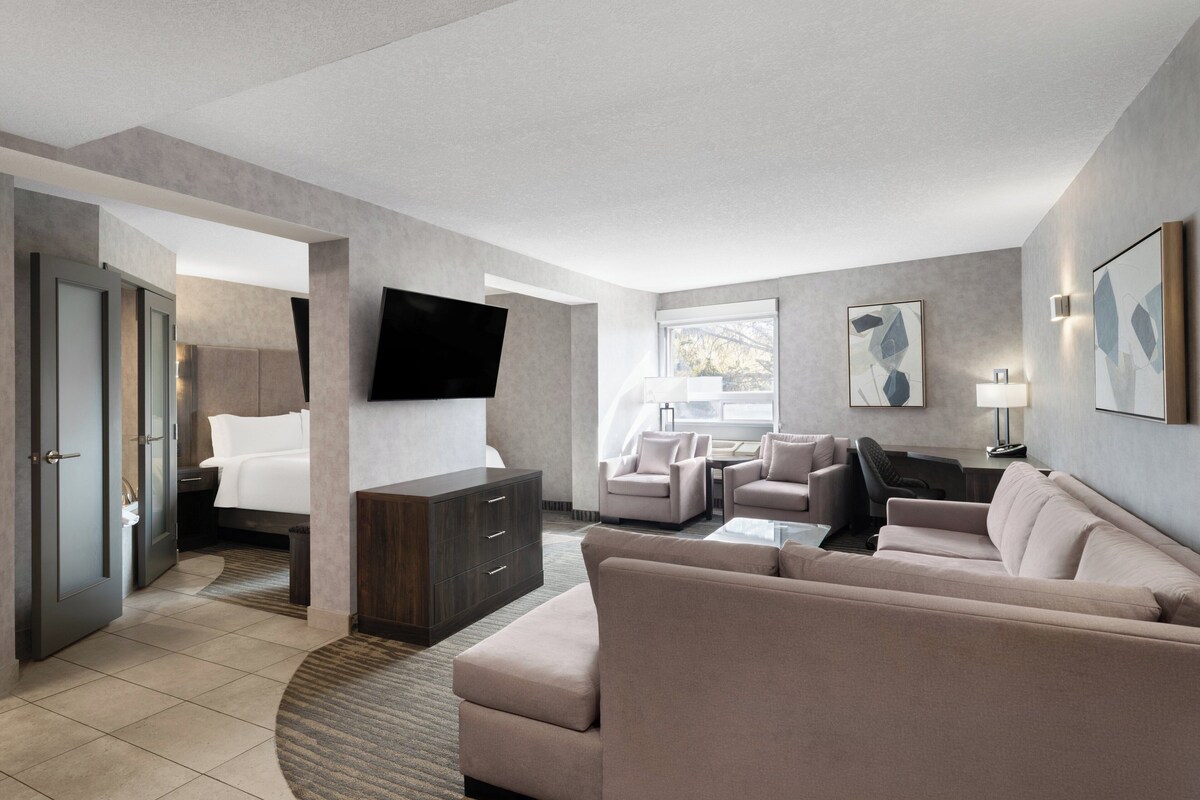 A modern hotel room with a large bed, a sofa, two armchairs, a flat-screen TV mounted on the wall, and a desk with a lamp.