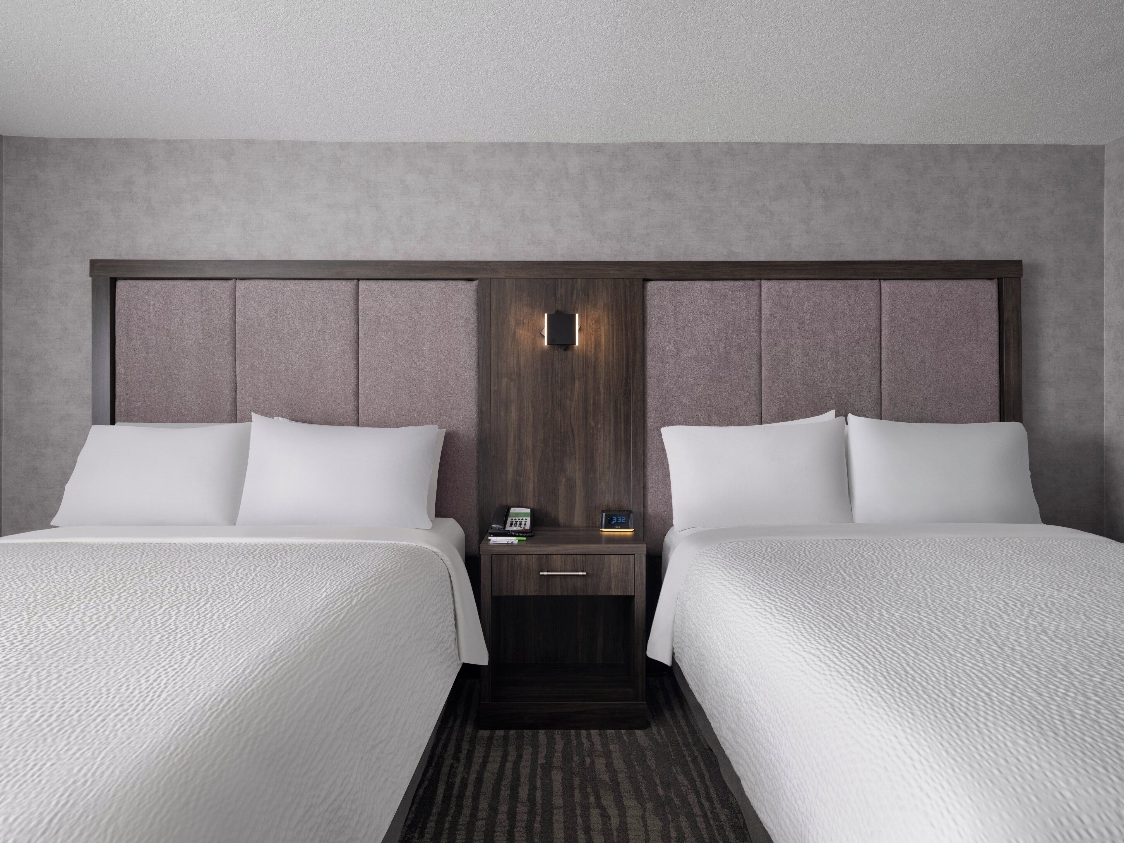 A hotel room with two beds, a wooden headboard, a nightstand with a phone and a clock, and a textured carpet.