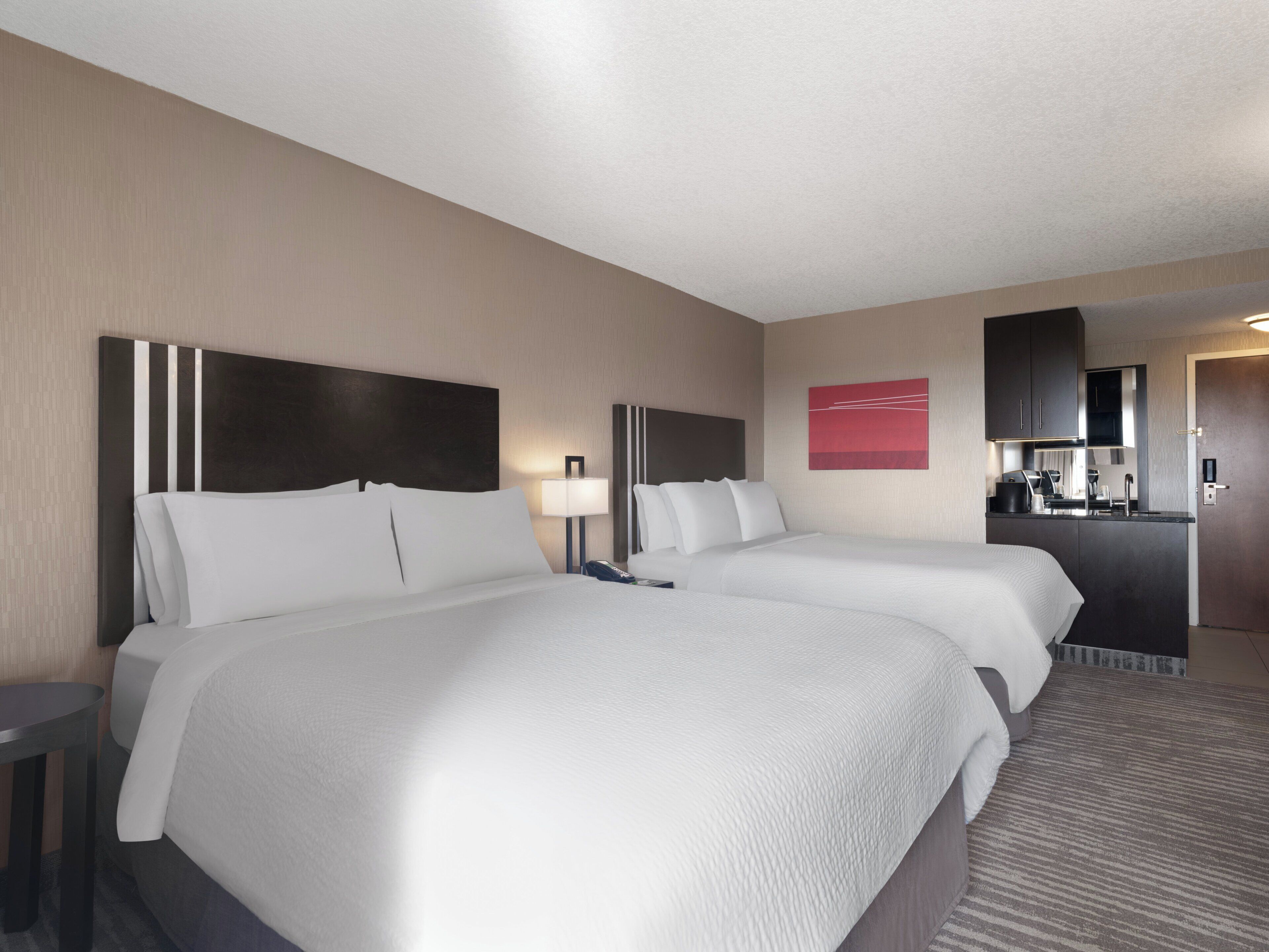 A hotel room with two beds, a desk, a lamp, a mirror, a red artwork, and a kitchenette.